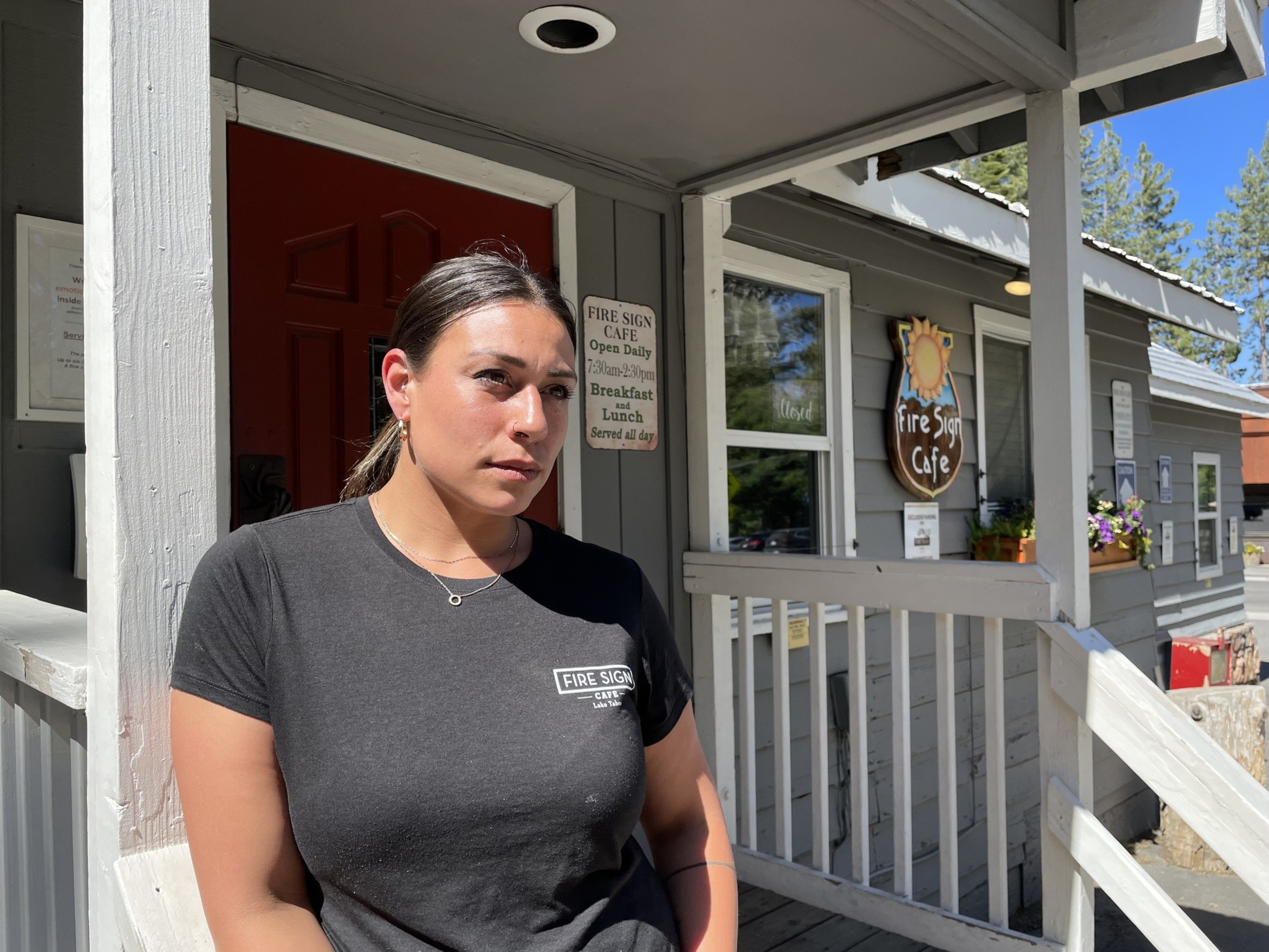 A woman stands in front of a gray building with a red door, porch, and flowers. She wears a black &quot;Fire Sign Café&quot; t-shirt. The café sign and some trees are visible.
