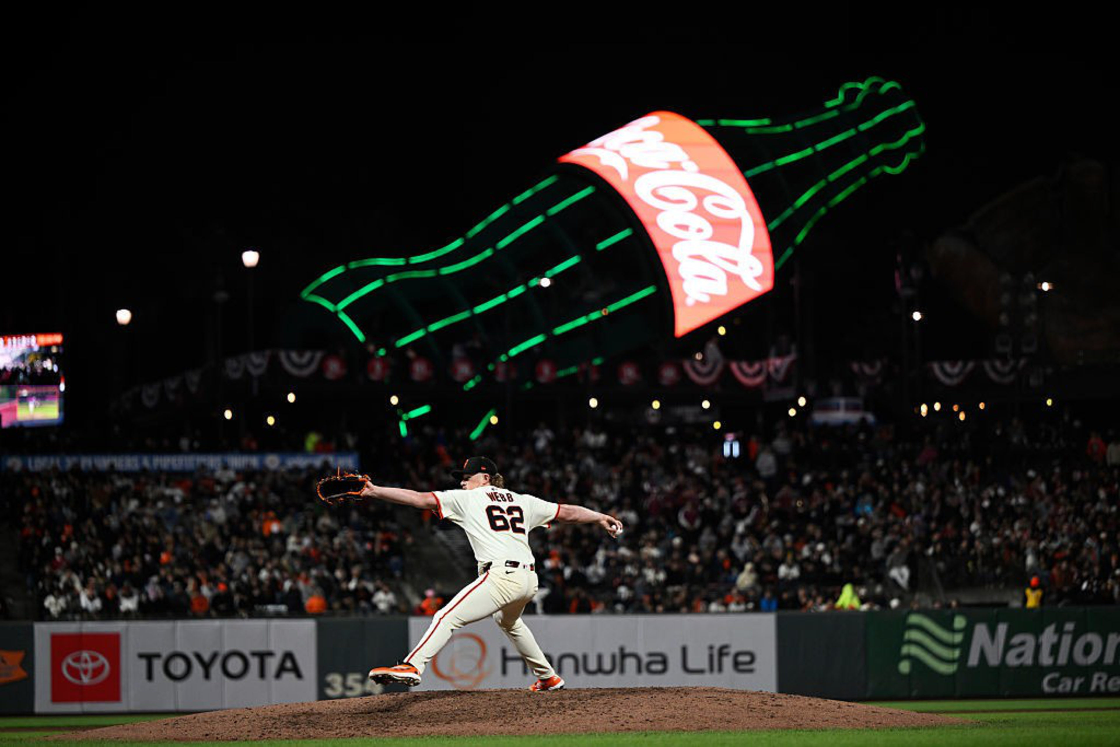 A baseball pitcher in a white uniform throws the ball under a large, illuminated Coca-Cola sign. The stadium is filled with spectators at night.