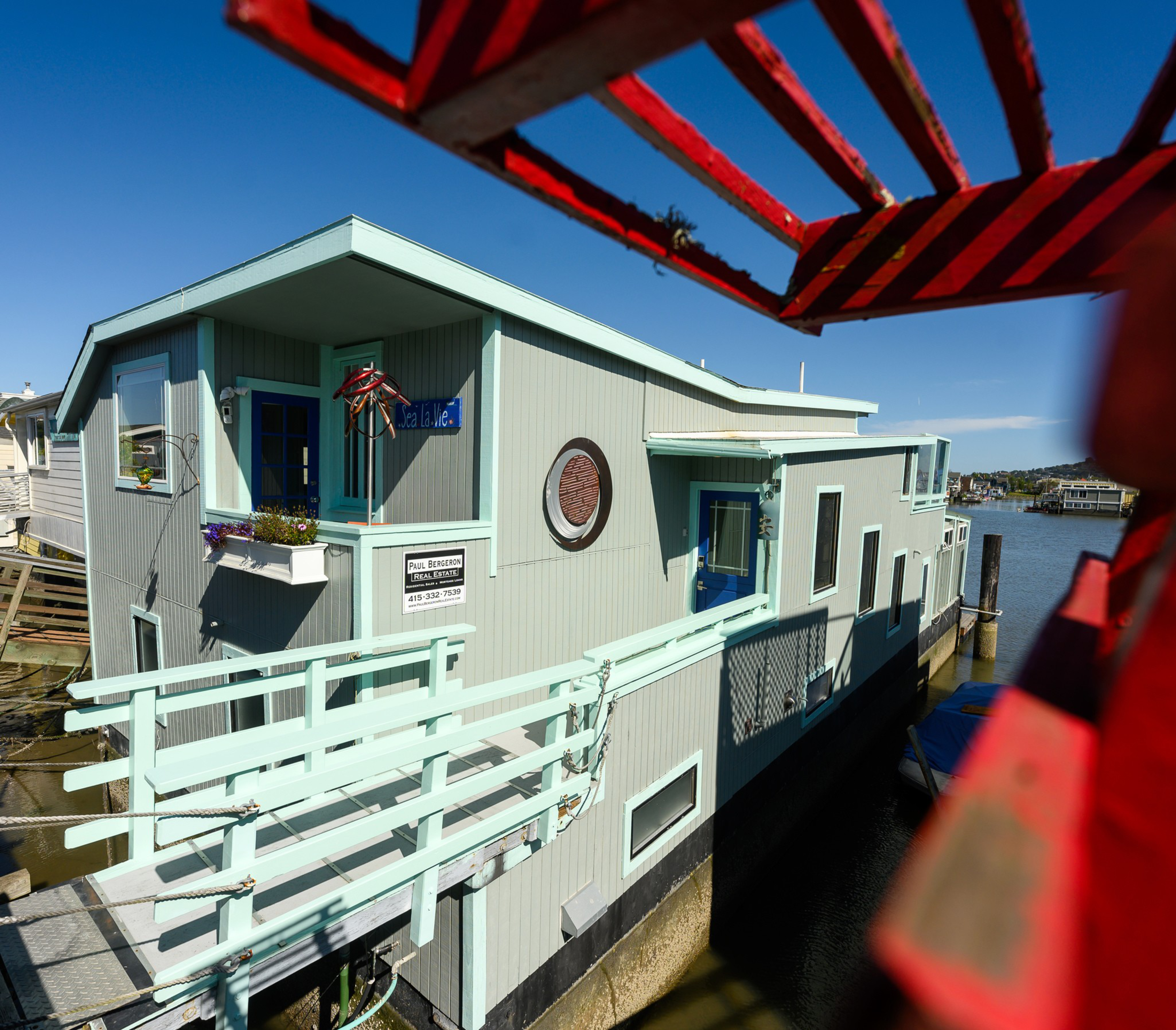 A floating house with a pale blue exterior, framed by red lattice, sits on calm water under a clear sky.