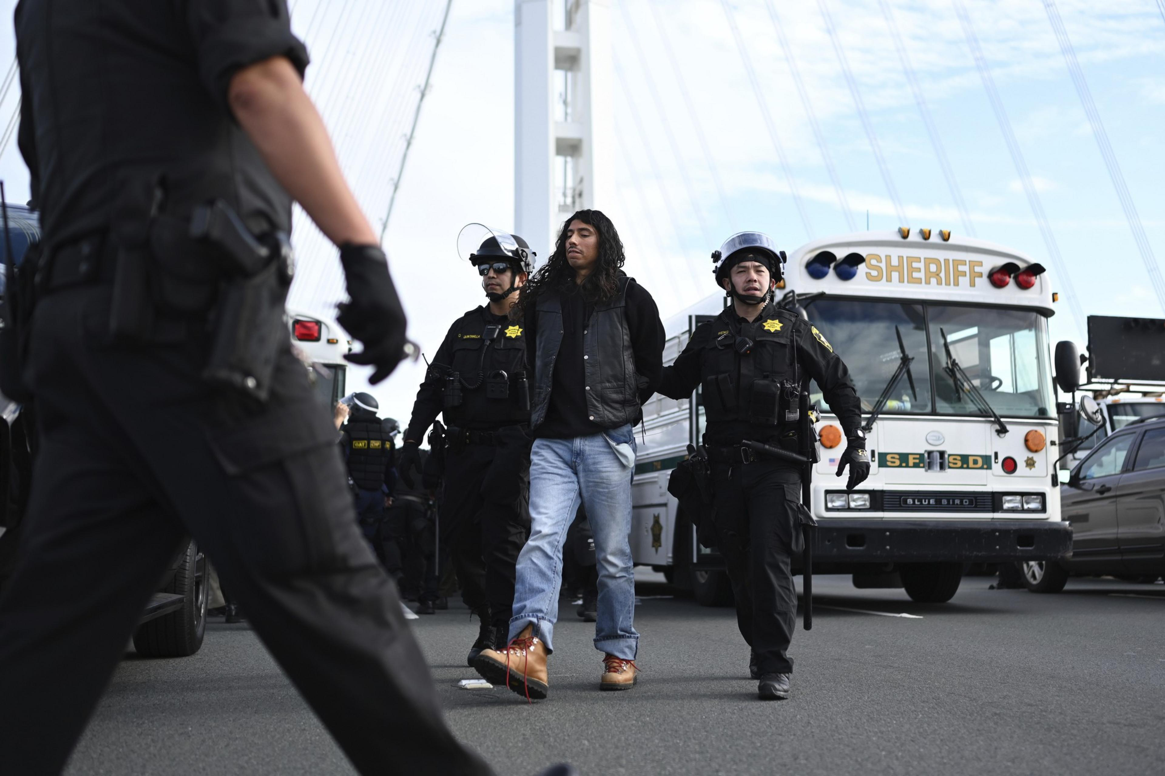 San Francisco Sheriff Department officers wearing riot gear escort a handcuffed person away with a bus that has "Sheriff" on the exterior.