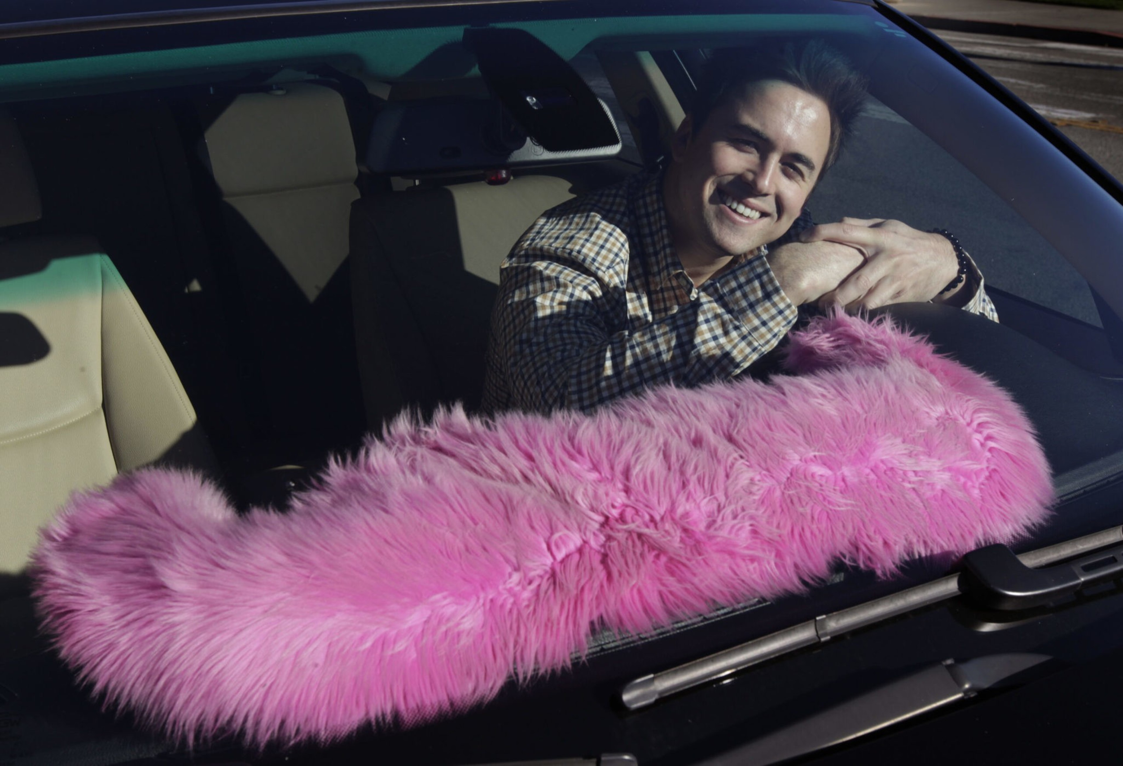A man with dark hair and a plaid shirt smiles while leaning on the steering wheel inside a car with a fluffy pink dashboard cover.