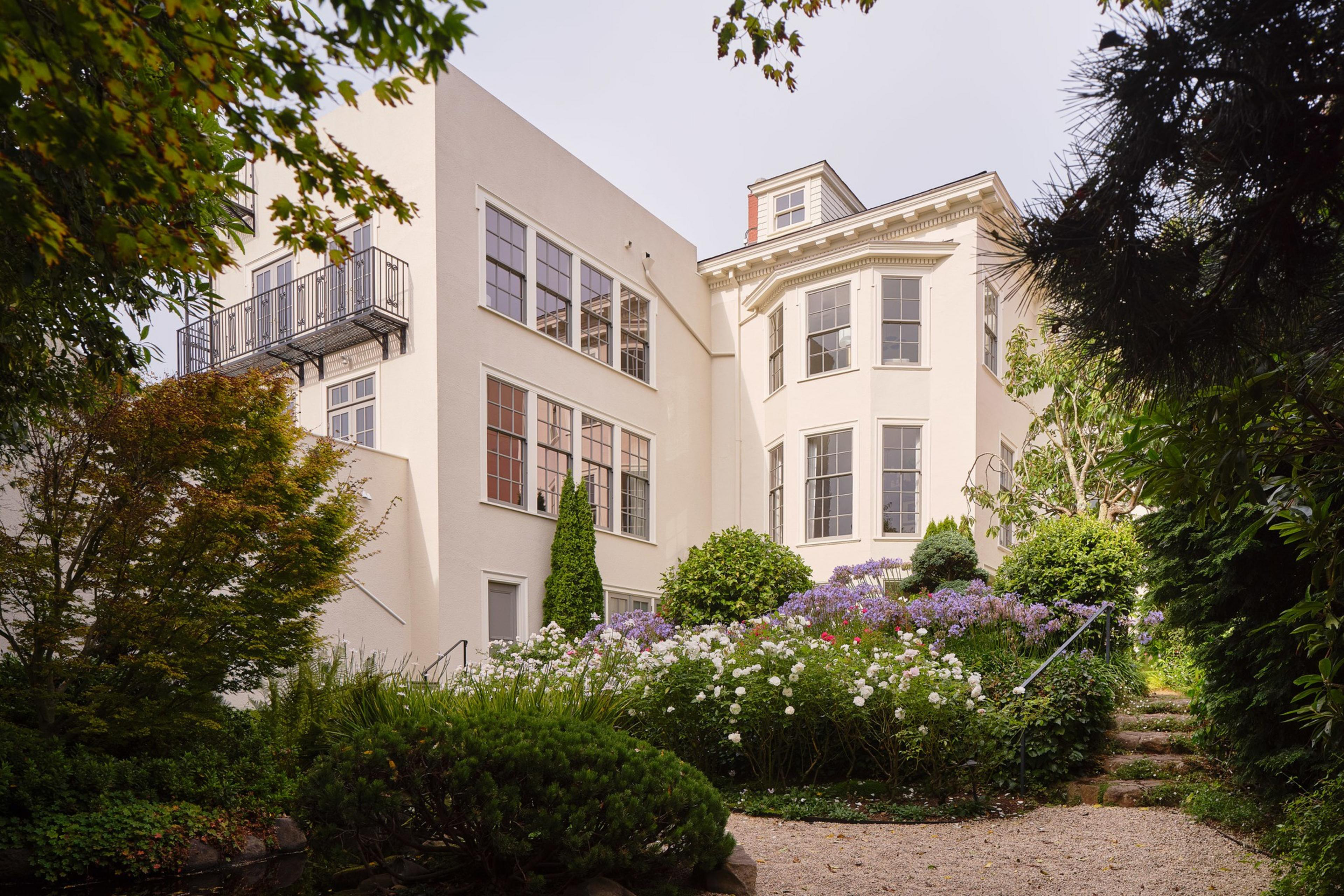 A large cream-colored house with multiple windows stands behind a lush garden filled with white, purple, and red flowers, surrounded by green bushes and trees.