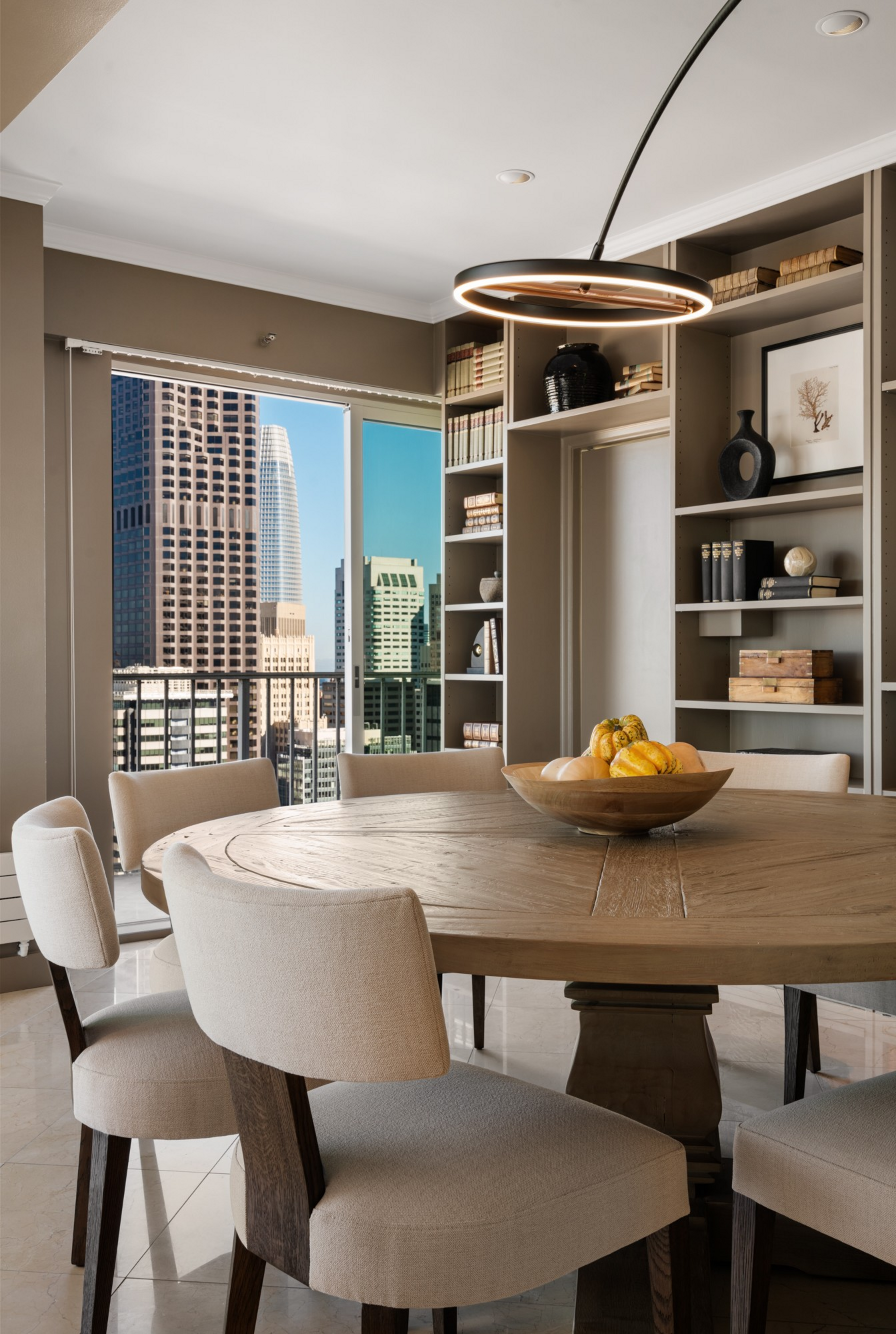 A round wooden dining table with six beige cushioned chairs surrounds a bowl of assorted gourds beneath a modern circular light fixture, with bookshelves and cityscape windows behind.