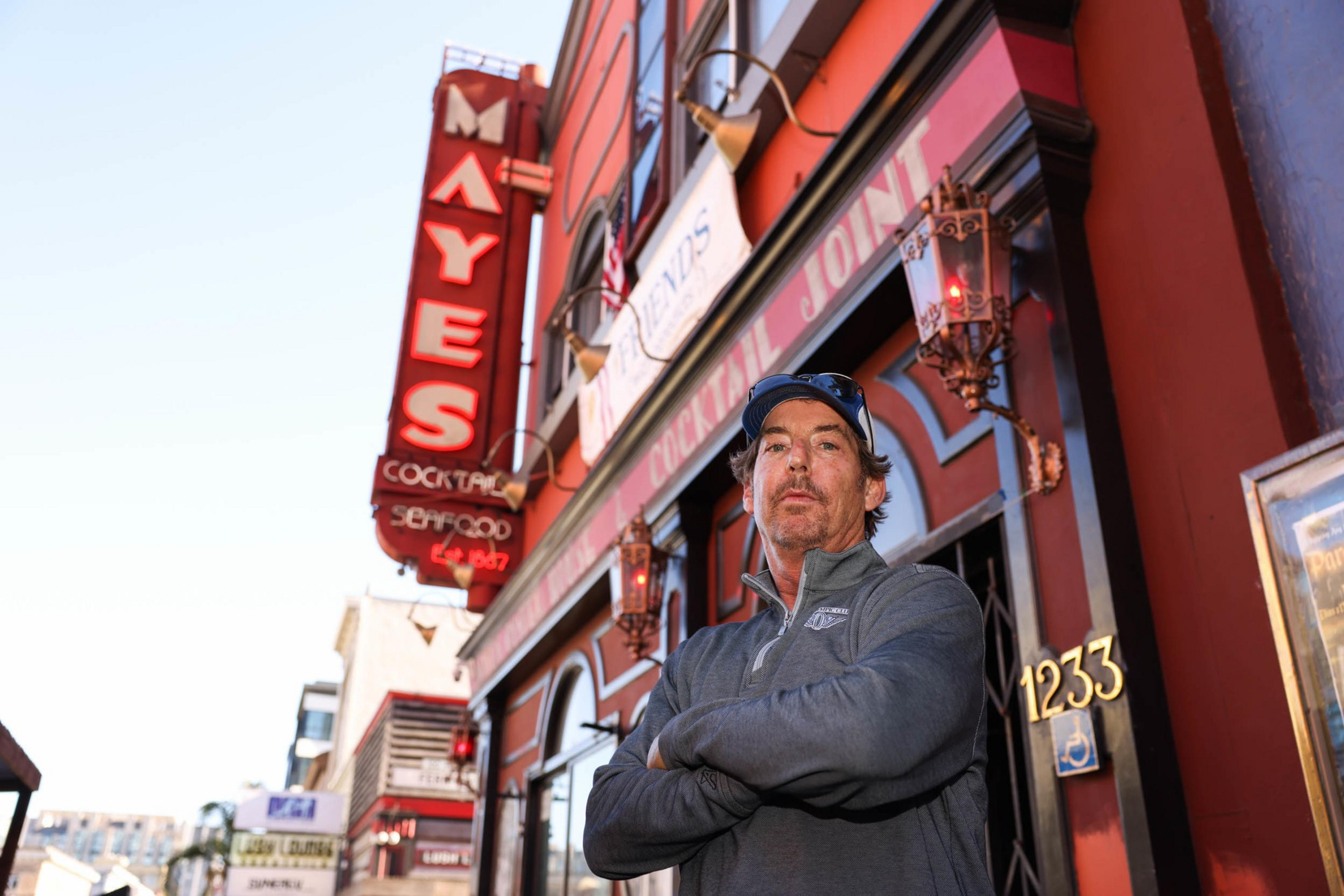 Matt Corvi man in a grey jacket stands with arms crossed in front of Mayes Oyster House restaurant, featuring a red facade and vintage neon sign.
