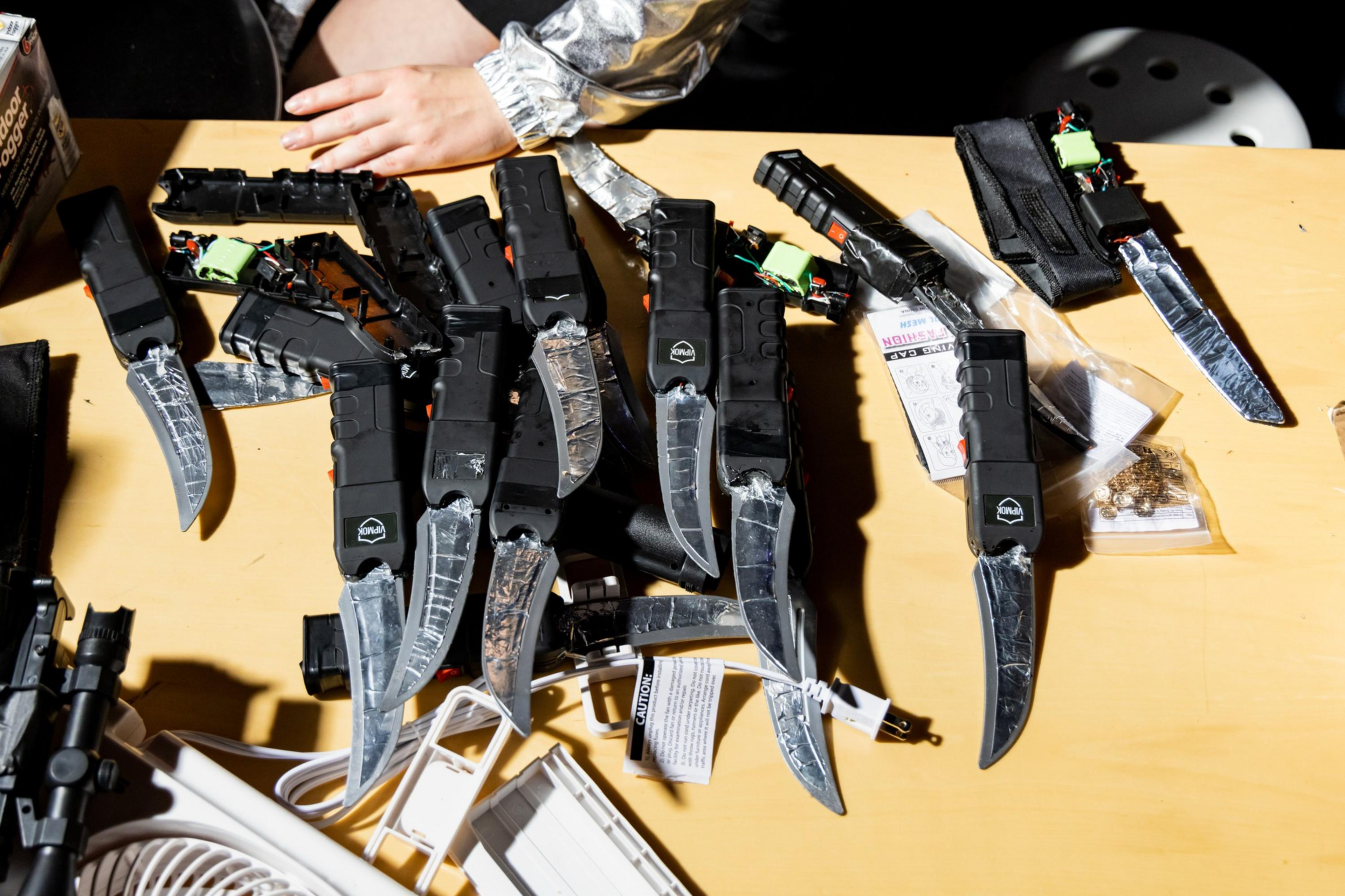 Several black knives with serrated and straight blades, some wrapped in tape, are spread out on a wooden table with a person's arm visible above.