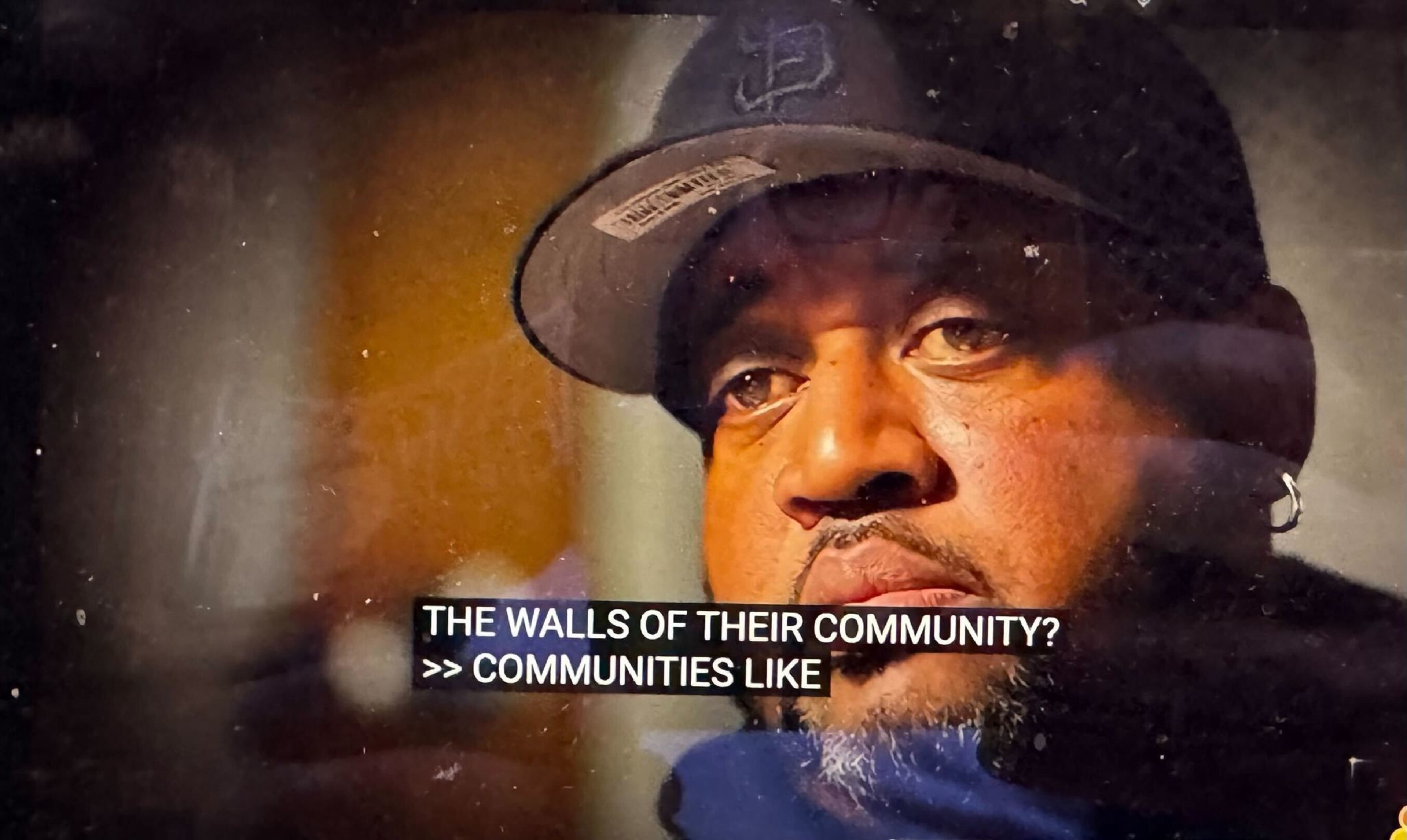 A close-up of a person wearing a hat and earrings with a serious expression. Subtitles read: &quot;THE WALLS OF THEIR COMMUNITY?&quot; and &quot;&gt;&gt; COMMUNITIES LIKE.&quot;