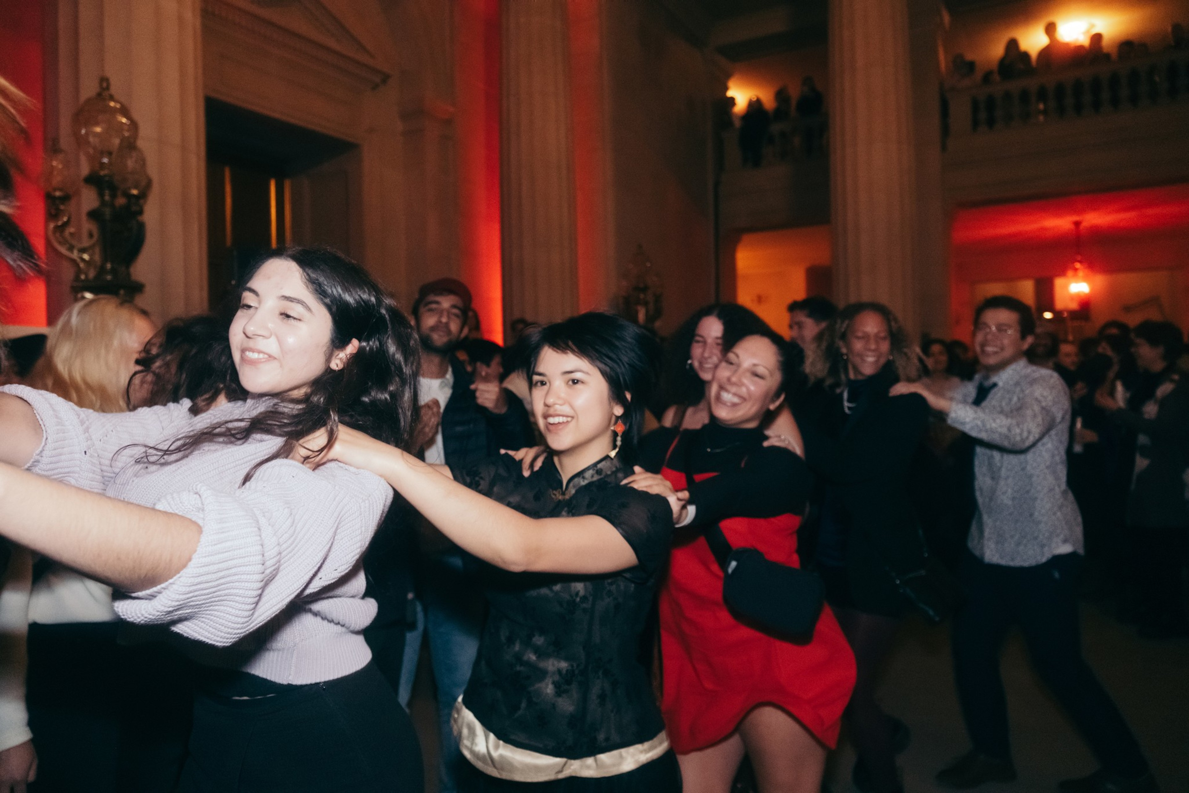 People dancing joyfully in a festive, dimly-lit hall with red lighting and classical architecture.