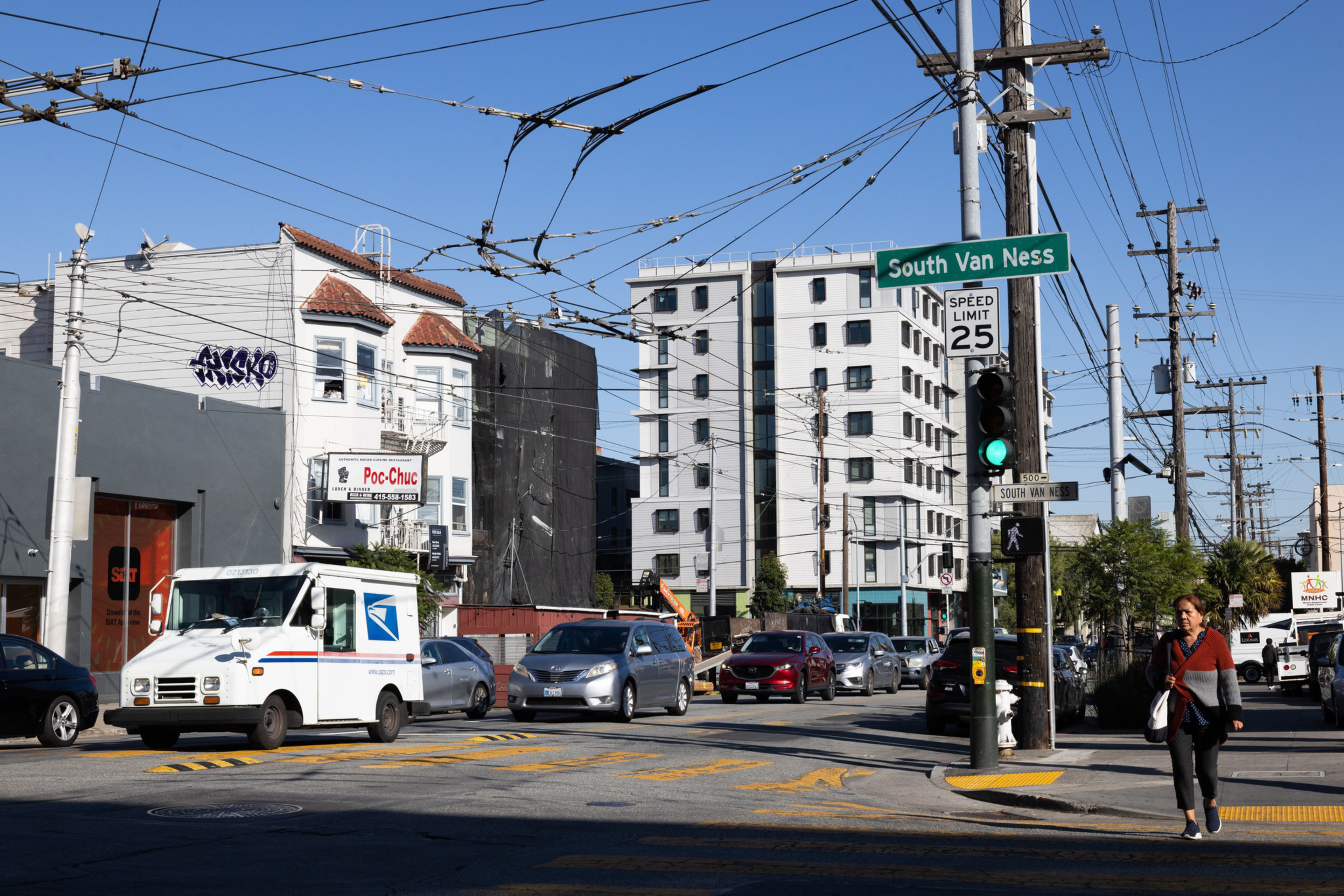 A busy urban intersection features cars, a mail truck, and a pedestrian crossing under a clear blue sky with visible power lines and buildings.