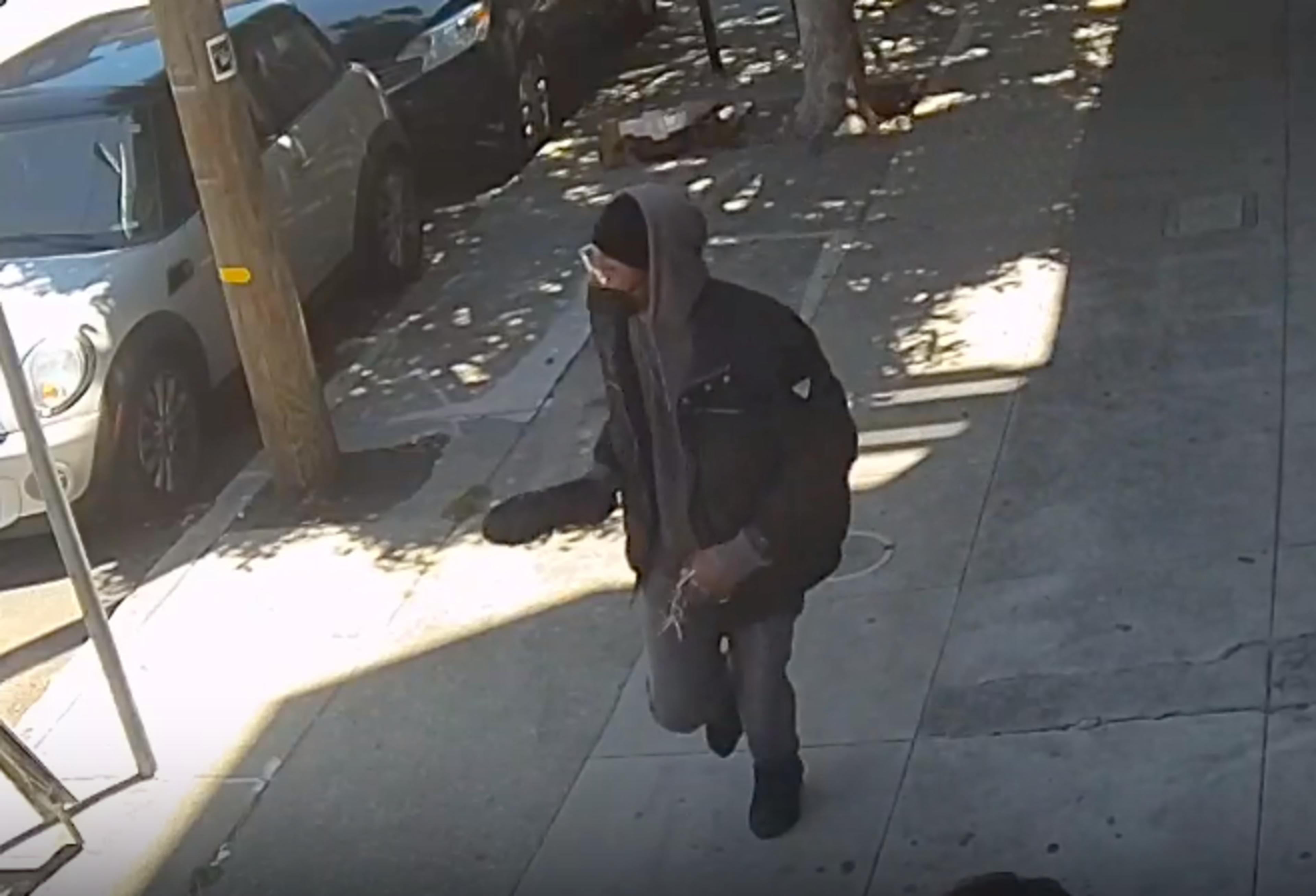 A person wearing a hooded jacket, face mask, and glasses walks on a sunlit sidewalk near parked cars and a utility pole.