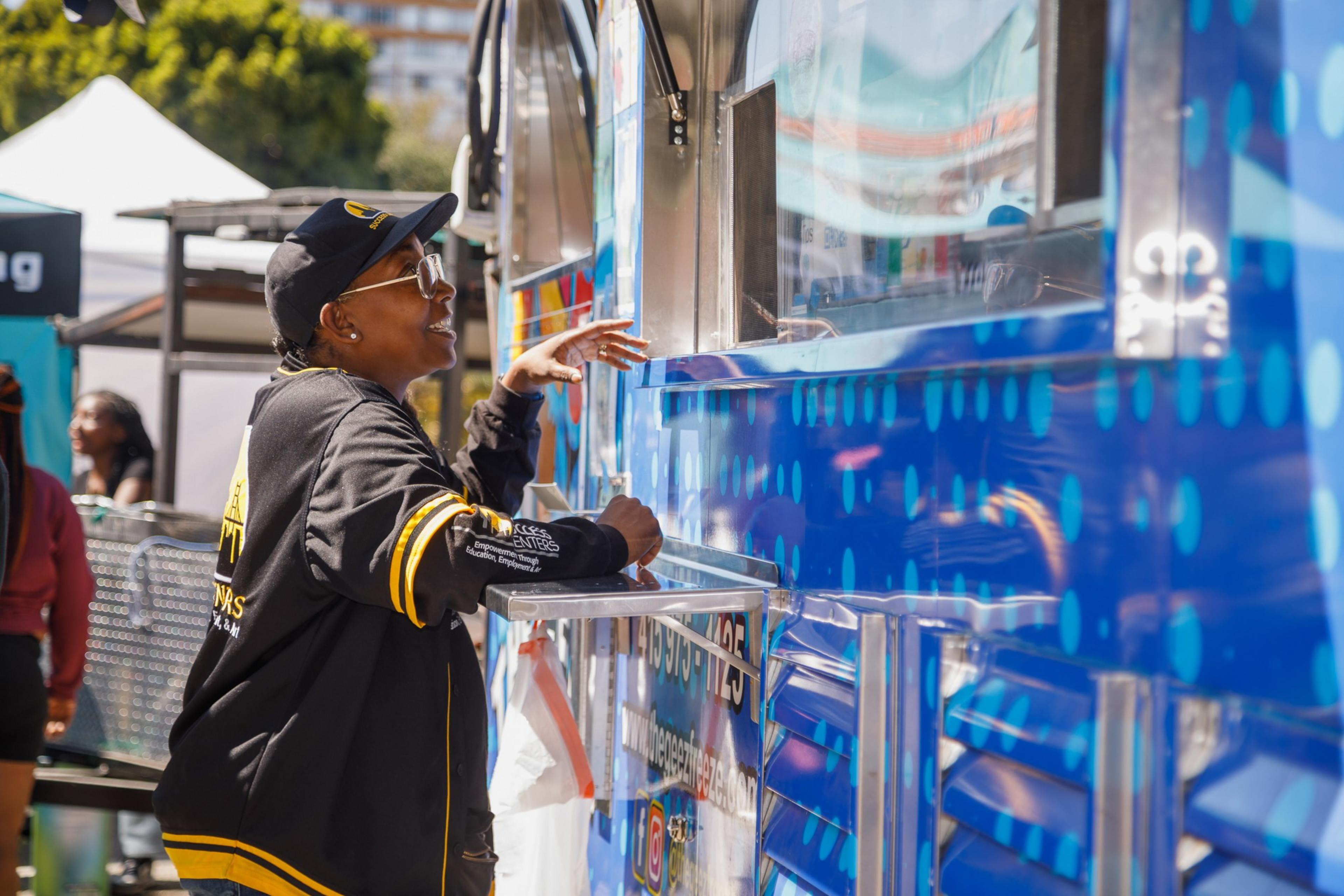 A person is ordering at a vibrant food truck on a sunny day, with other people around.