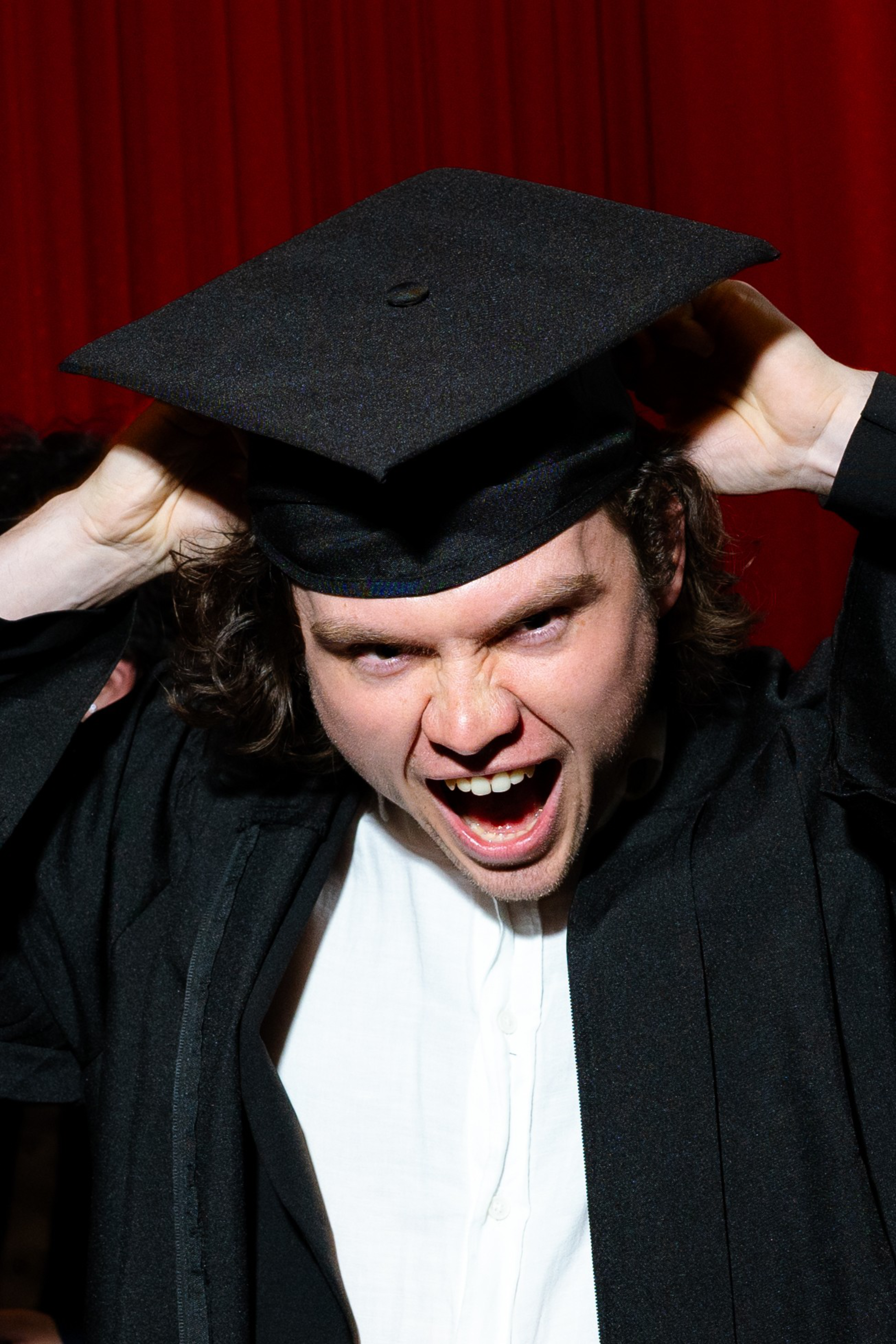 A person is wearing a graduation cap and gown, making an excited expression with their mouth open wide, in front of a red curtain.