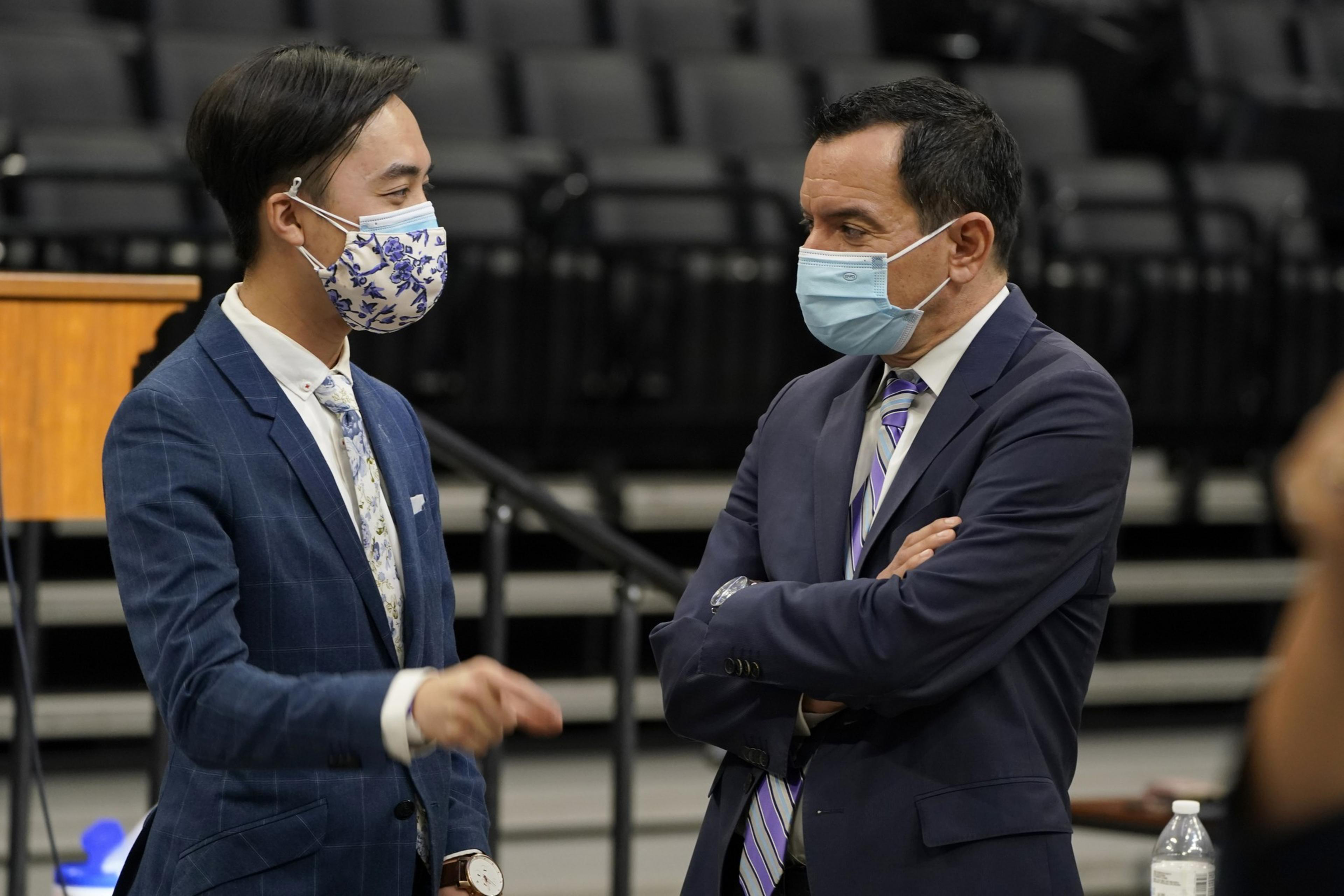 Two men in suits and masks face each other, seemingly in conversation, with empty seats in the background.