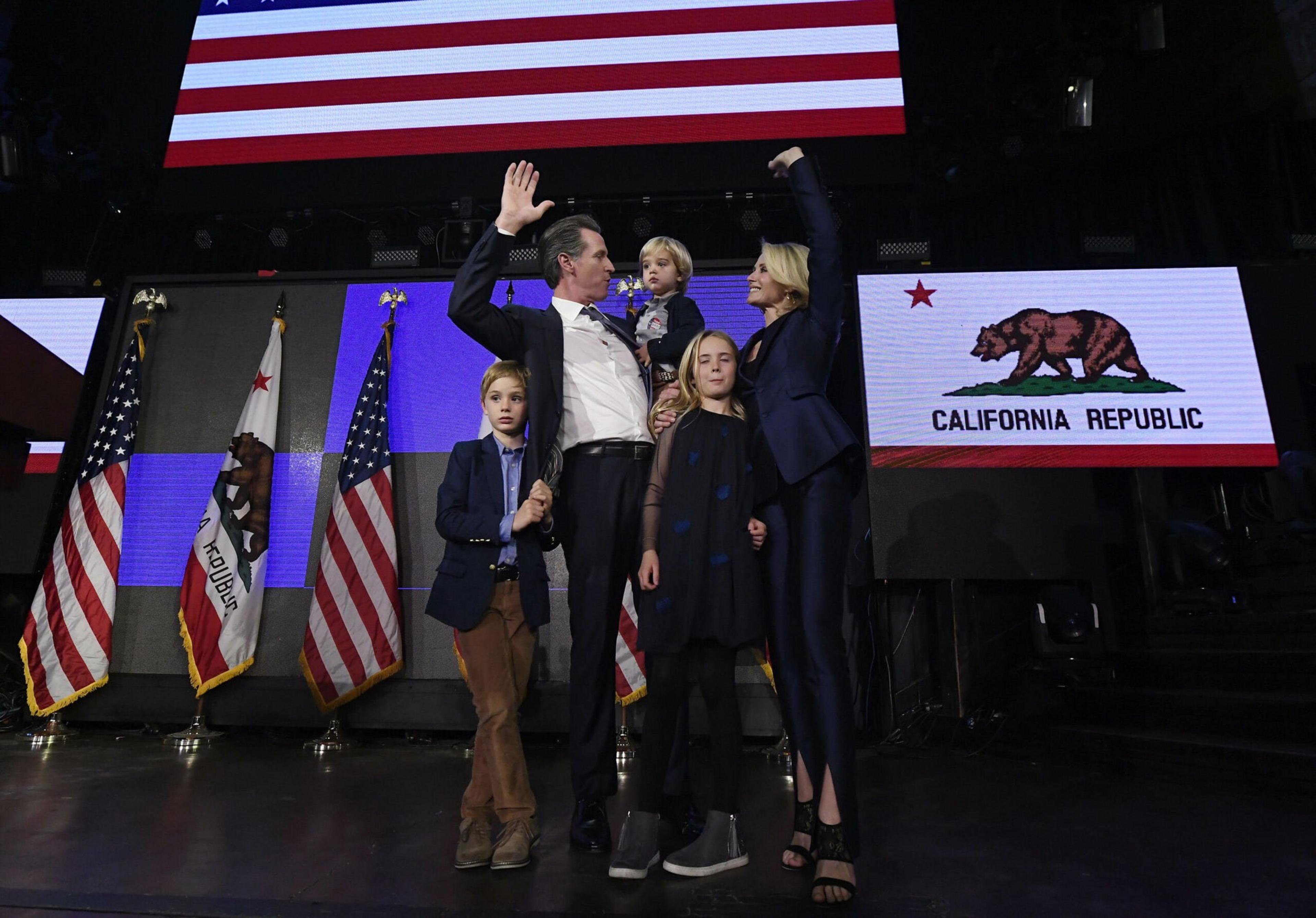 A family of five stands on stage with American and California flags behind them, with the father and mother raising their hands in a celebratory gesture.