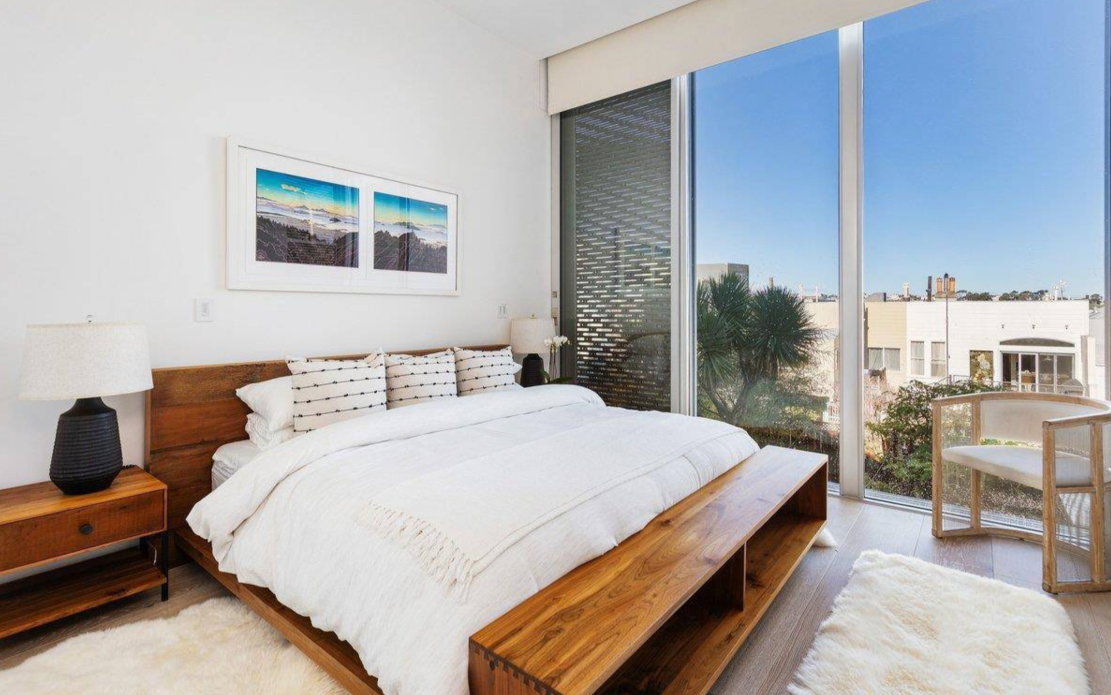 A modern bedroom features a wooden bed with white bedding, a matching bench, bedside tables with lamps, a large window with city views, and two white rugs.