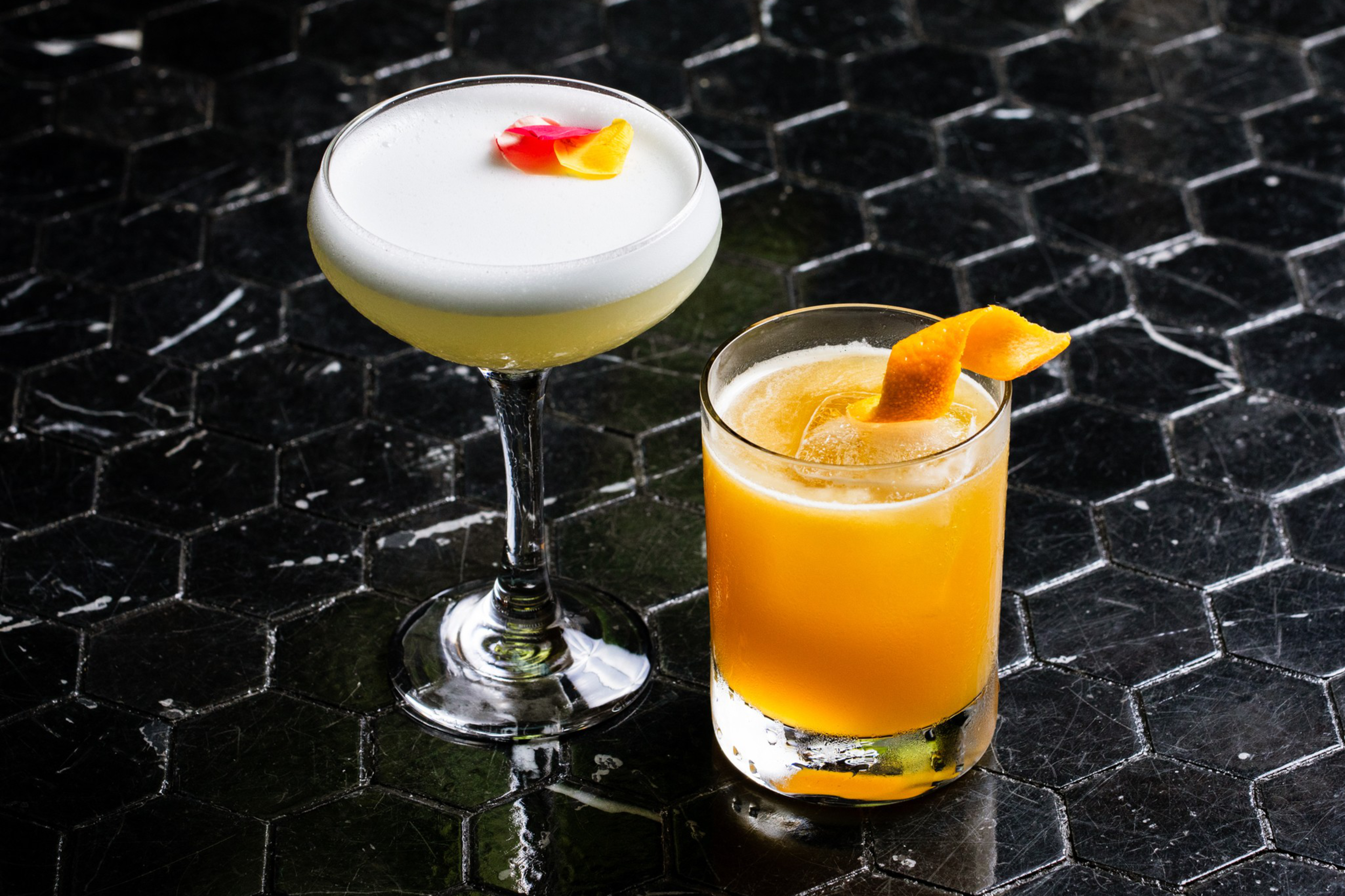 Two cocktails sit on a black hexagonal tile surface: one with a foamy top adorned with flower petals and the other an orange drink garnished with a citrus twist.