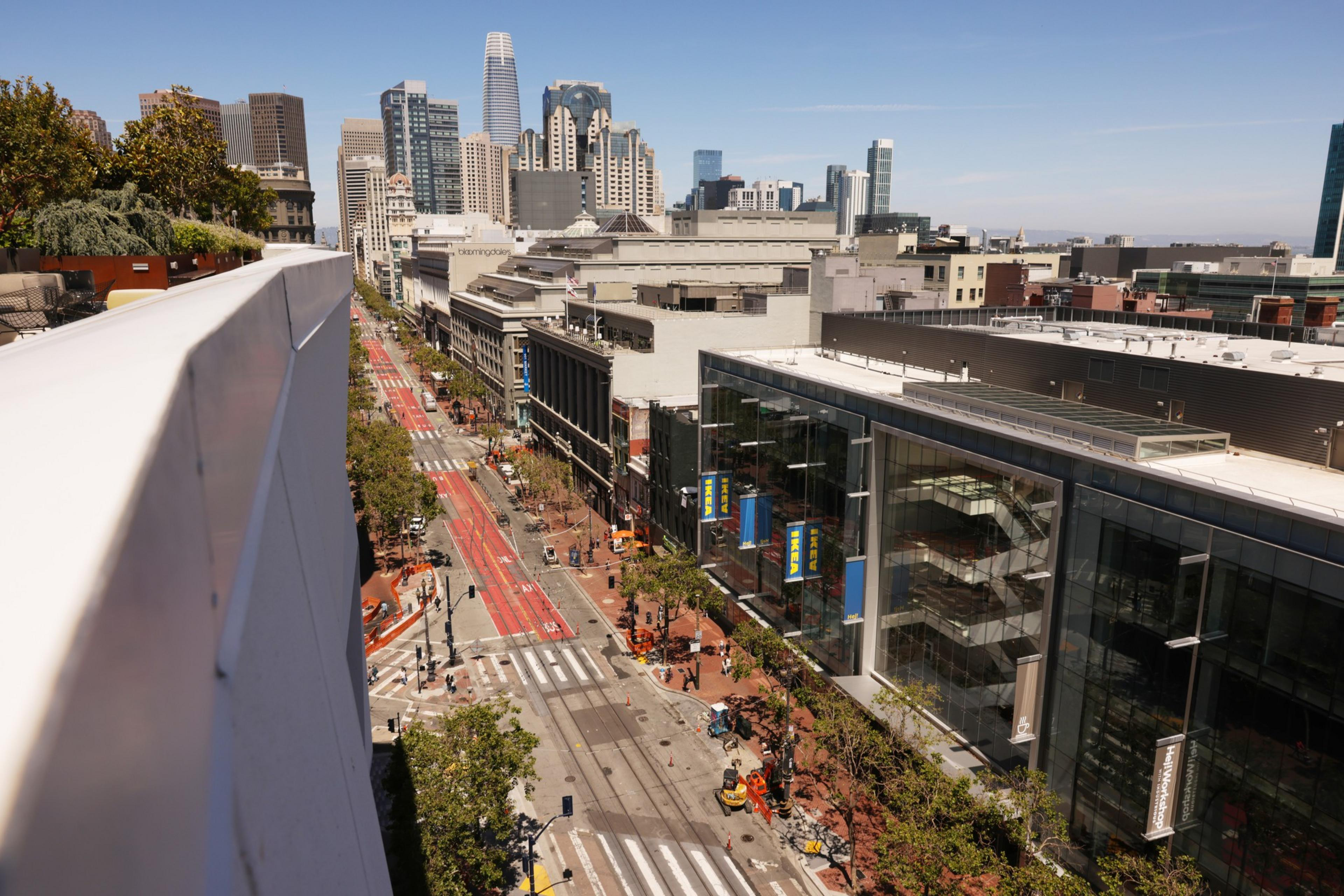 This image shows a busy urban street lined with buildings, trees, and red bus lanes, with skyscrapers in the background under a clear blue sky.