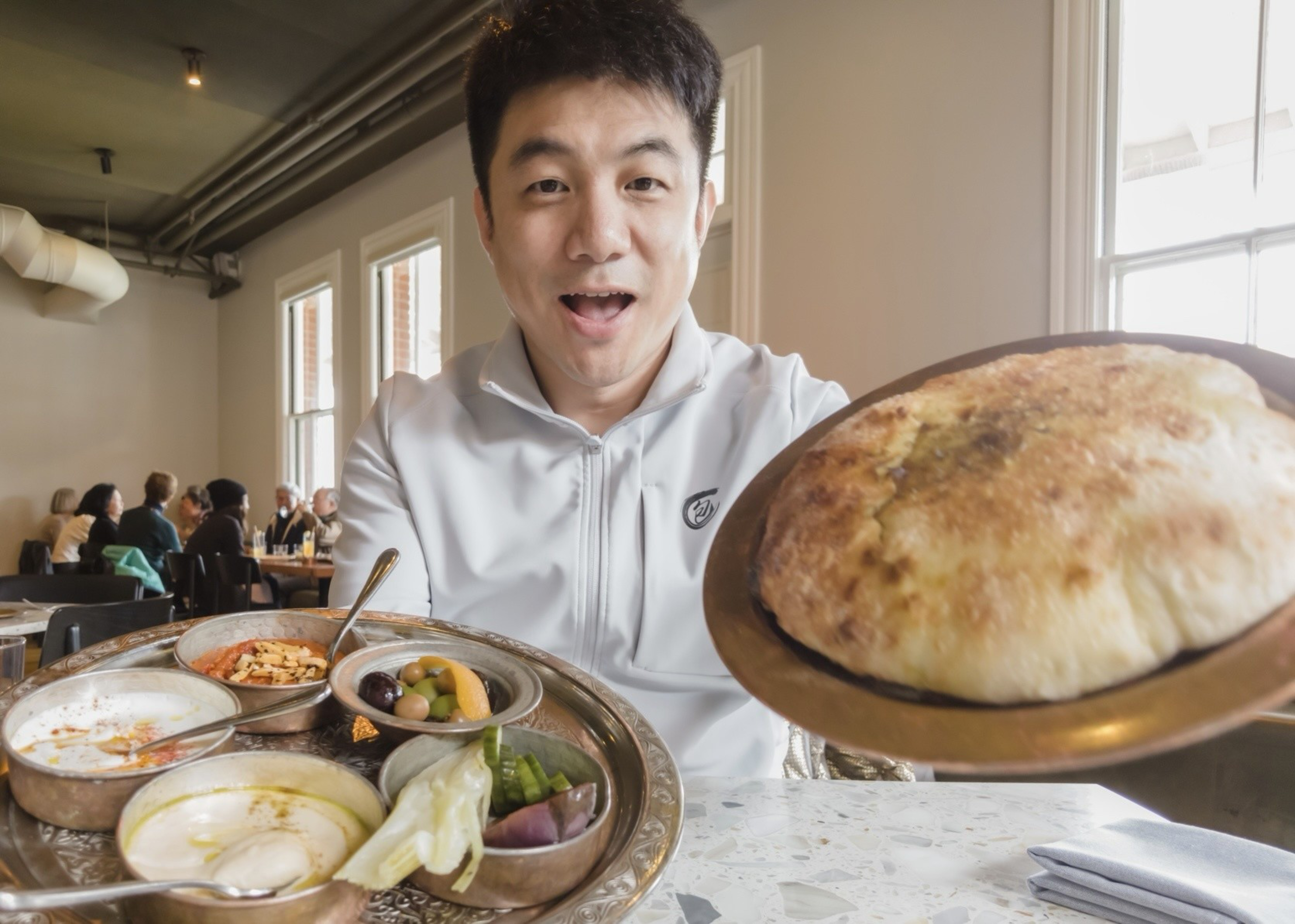 A man in a white jacket smiles, presenting a large, puffed bread and a tray of various colorful dishes in a busy restaurant.