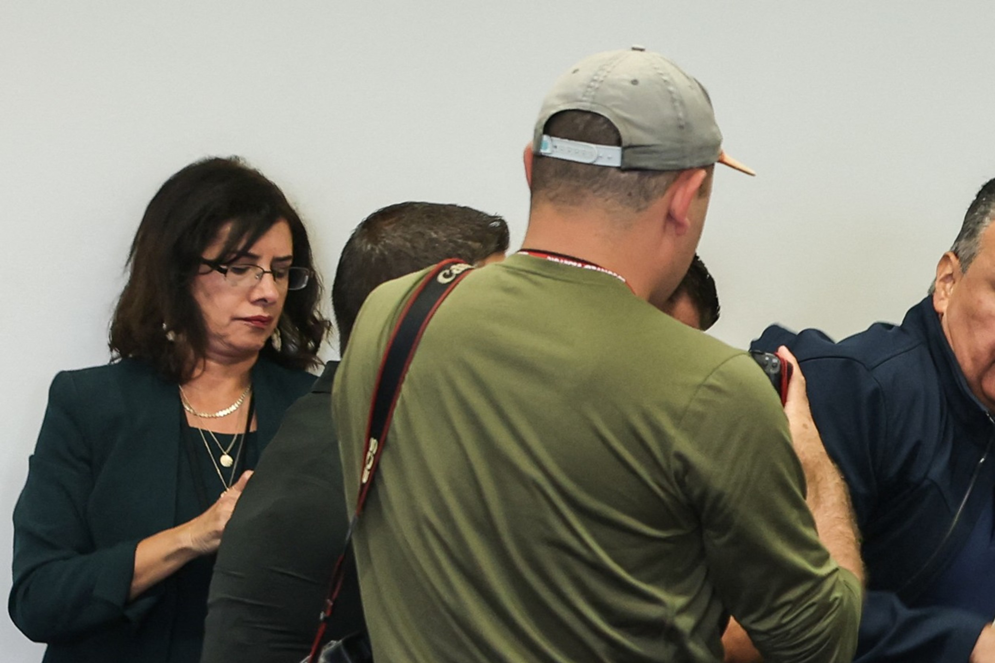 A media scrum captures a man in a navy sweater as he moves past a group, including a photographer in a cap with a camera and another person in a green jacket.