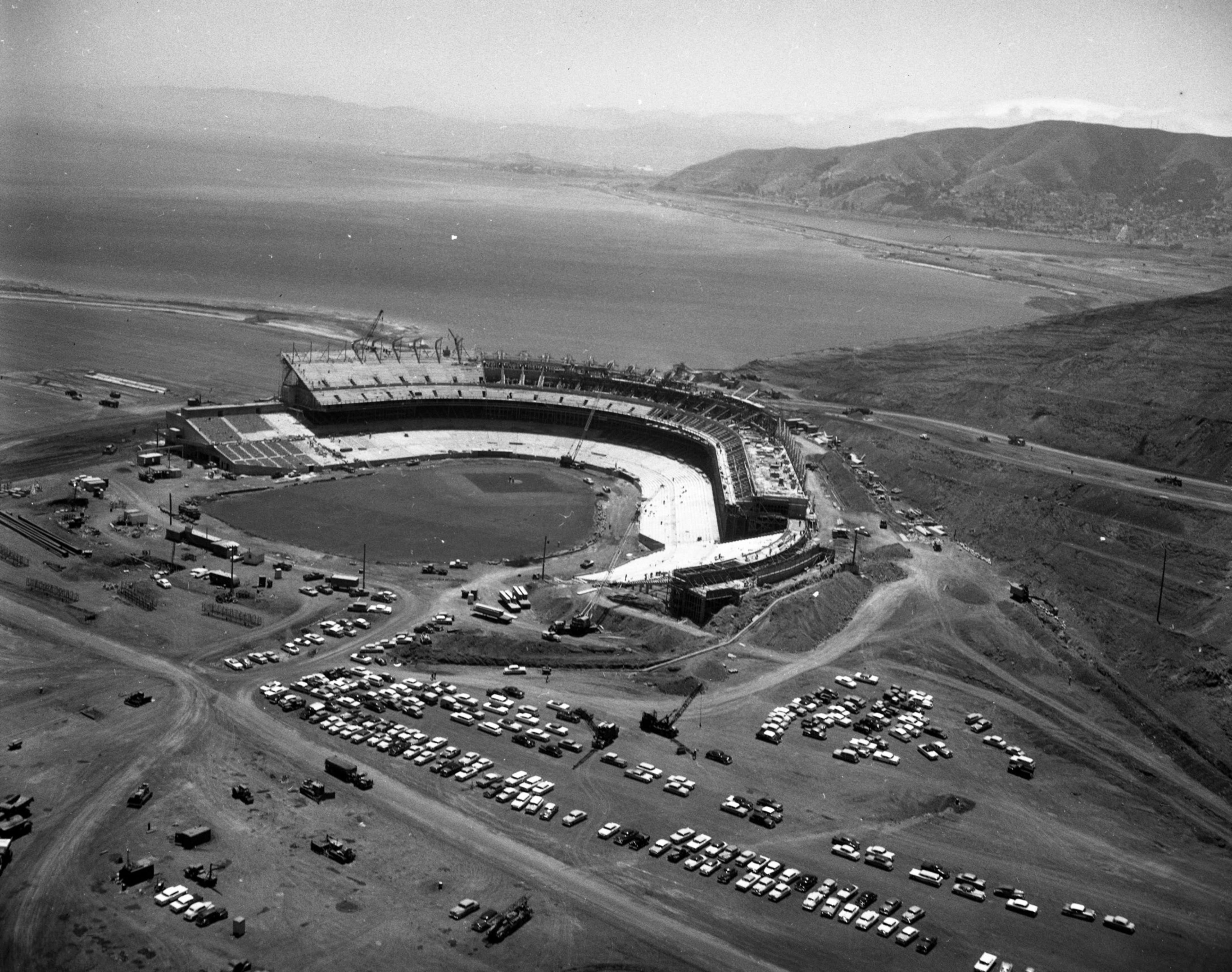 An aerial view shows a large, partially constructed stadium surrounded by dirt roads and parked cars, with a body of water and hills in the background.