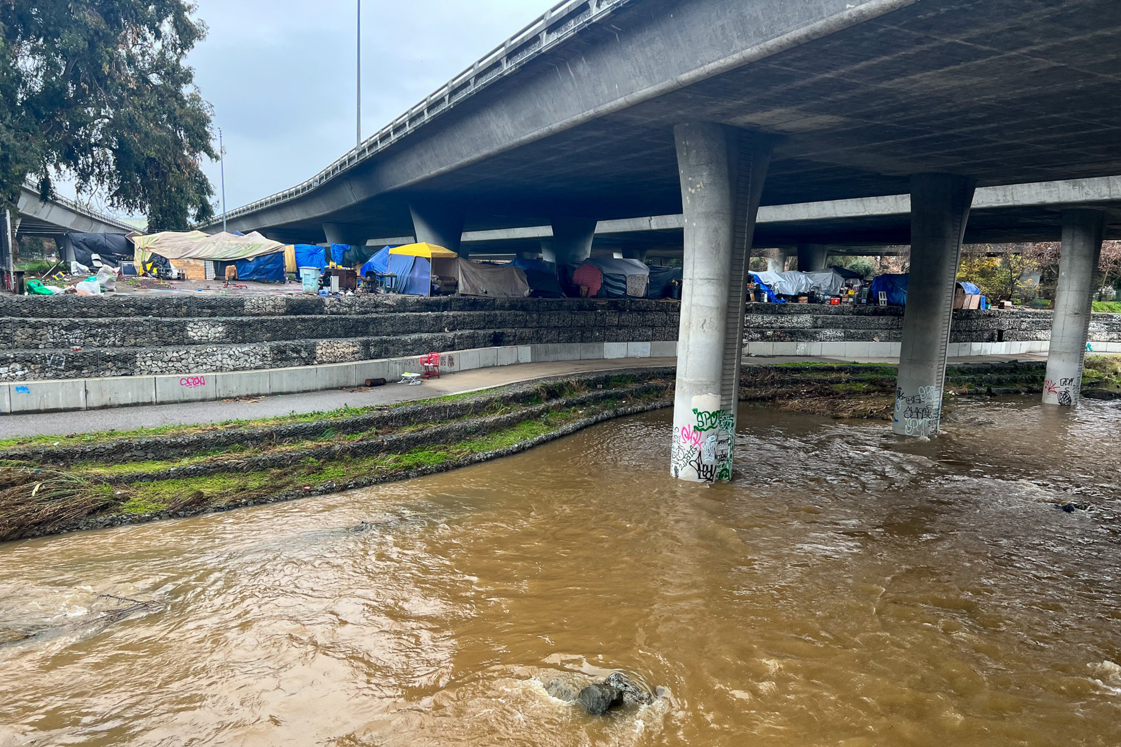A muddy river flows beneath a graffiti-covered concrete bridge, with tents and makeshift shelters set up along the riverbank.
