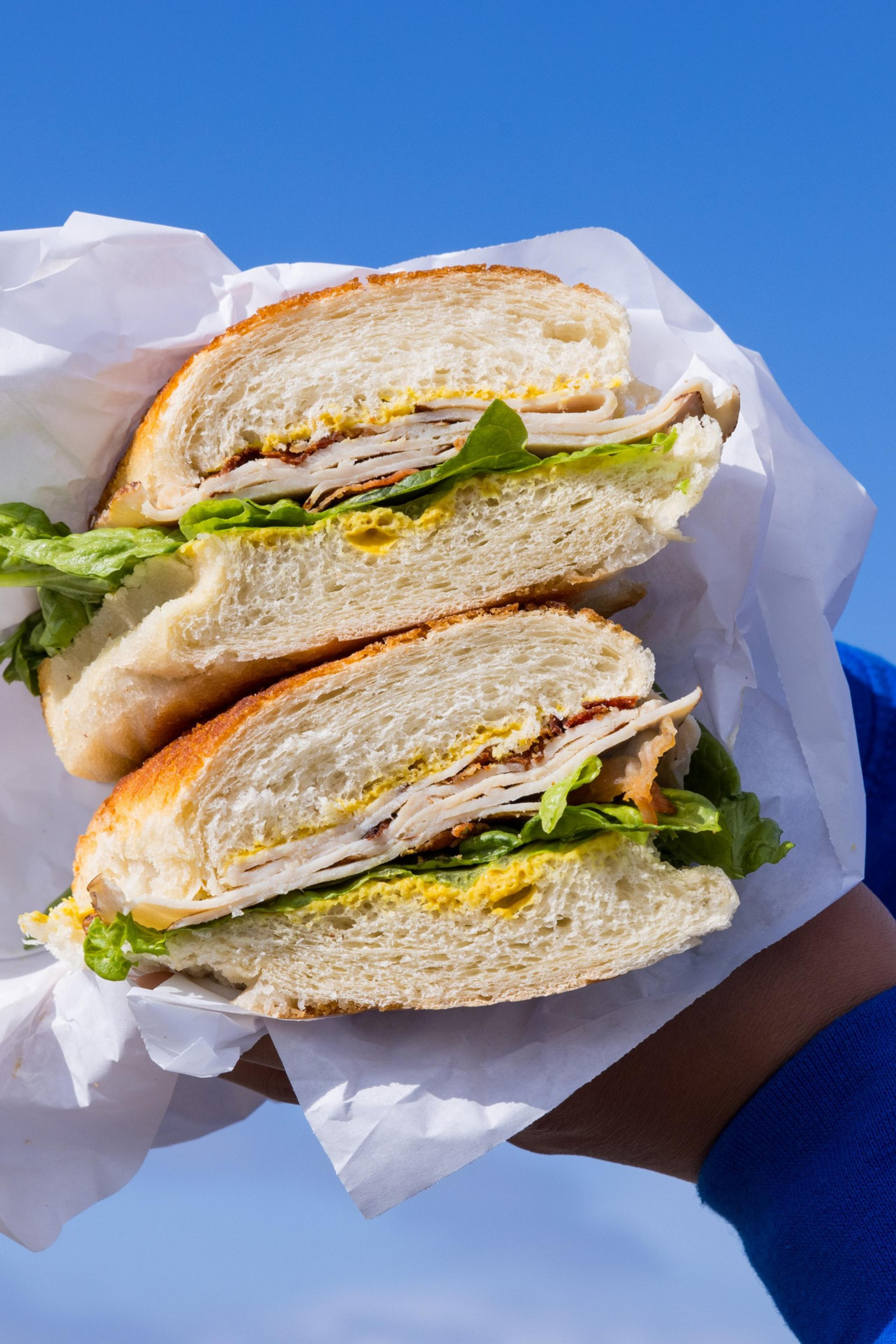 A person holds a halved sandwich with lettuce, turkey slices, and mustard between white bread, wrapped in parchment paper, against a clear blue sky.