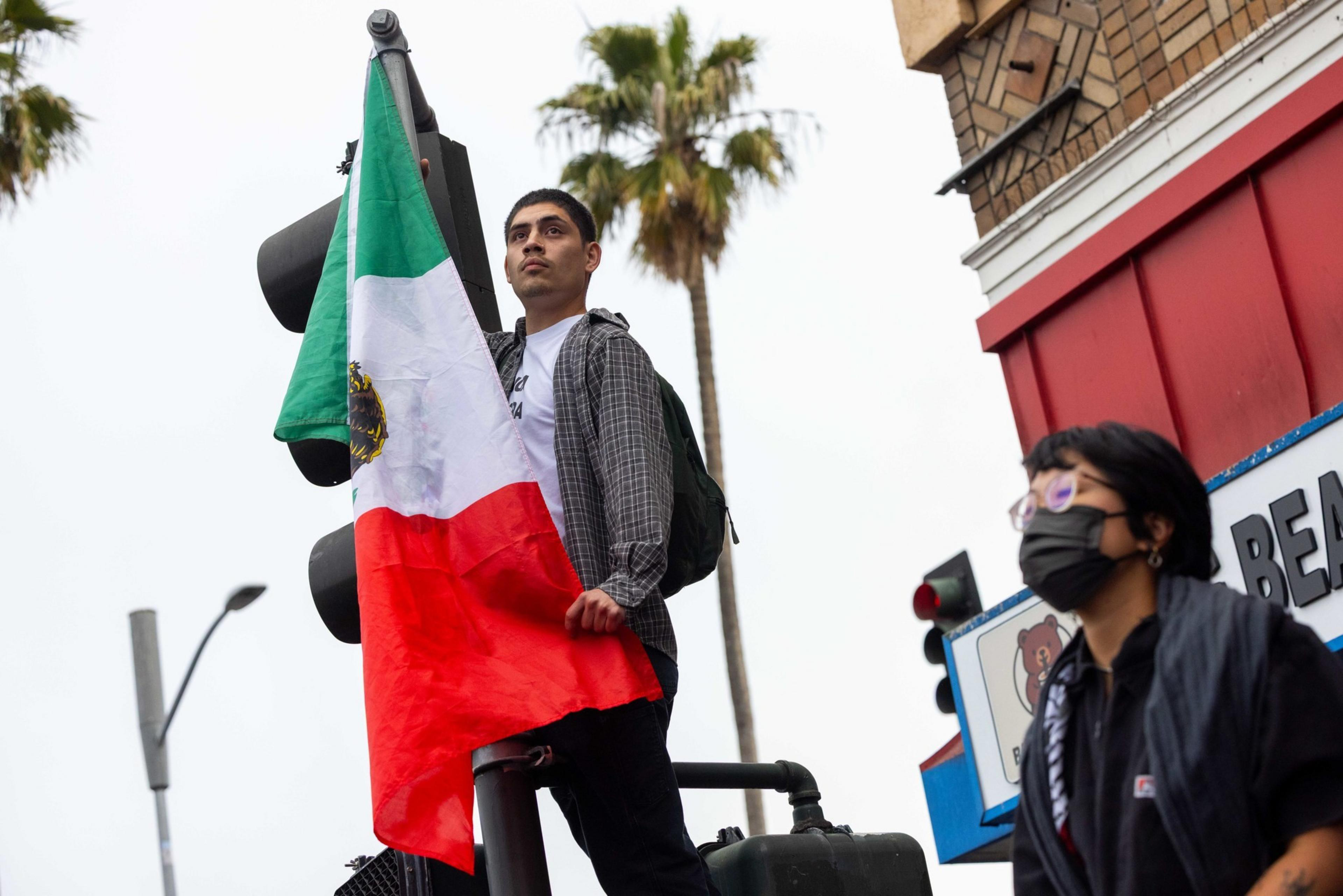 A person stands on a traffic light holding a Mexican flag. Another person in a mask walks nearby. Palm trees and a building are in the background.