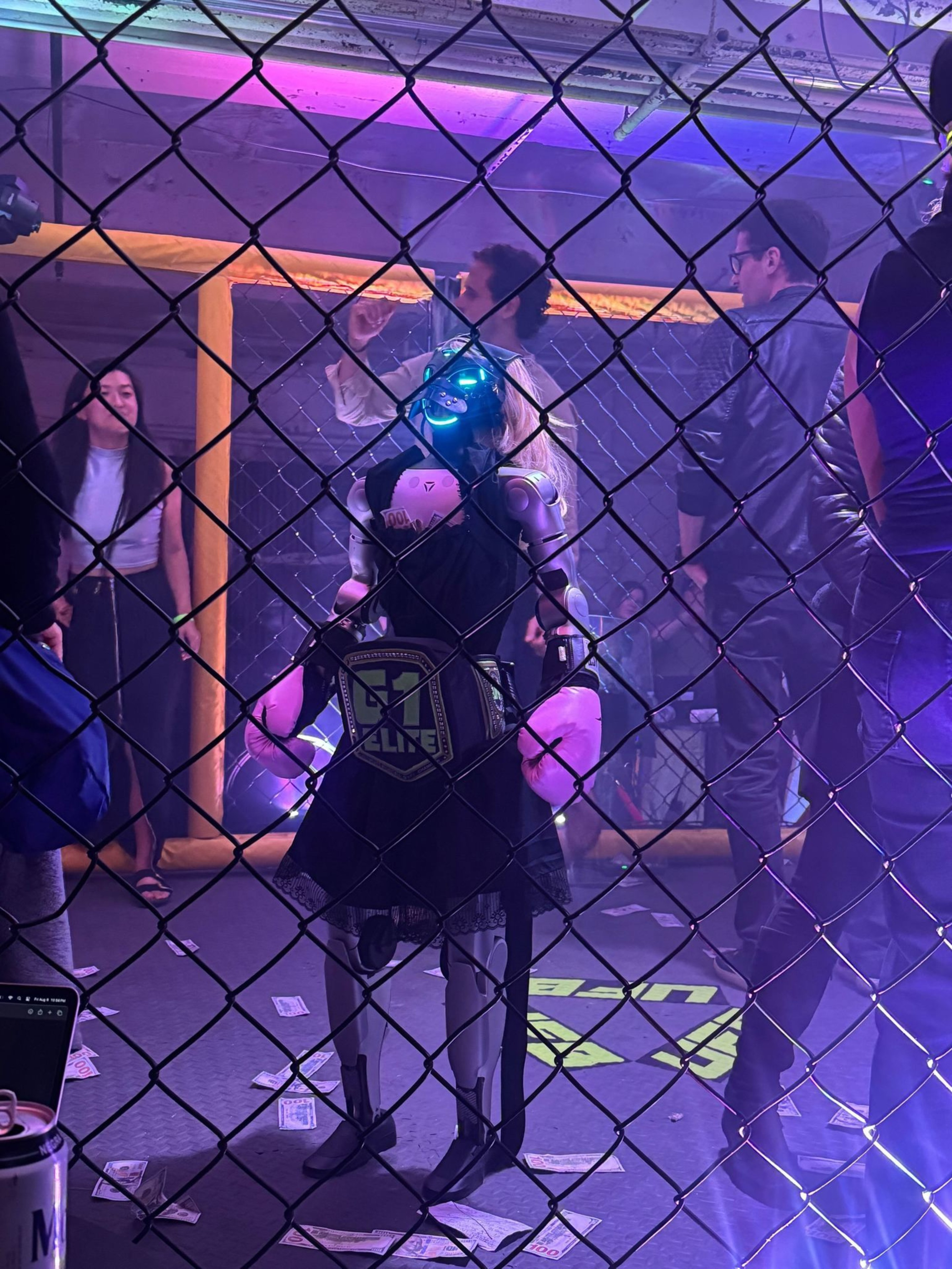 A robot wearing a skirt and boxing gloves stands inside a cage ring with people around, under purple and blue lighting.