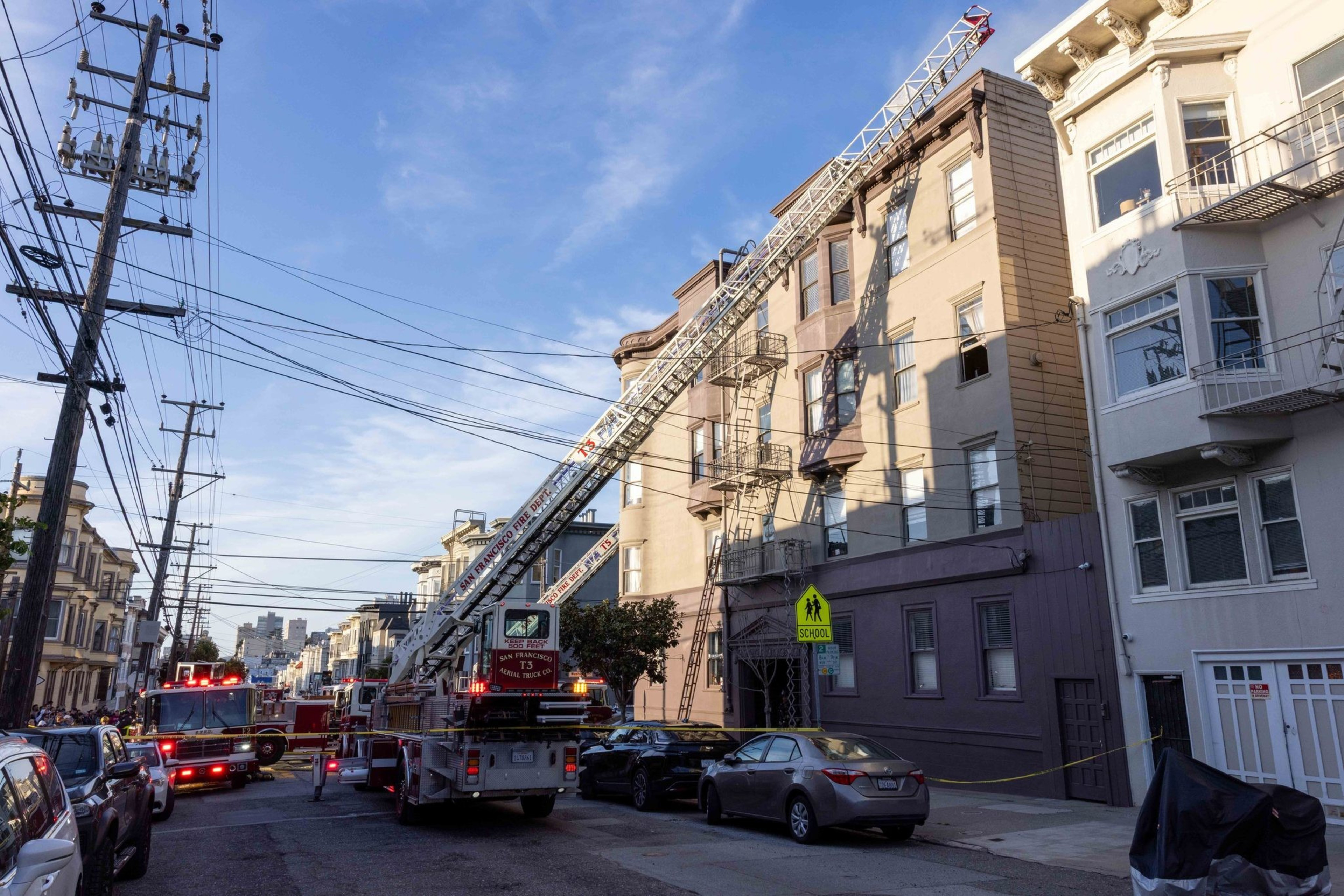 A fire truck with an extended ladder is positioned near a multi-story building. Several emergency vehicles and personnel are present on the street.