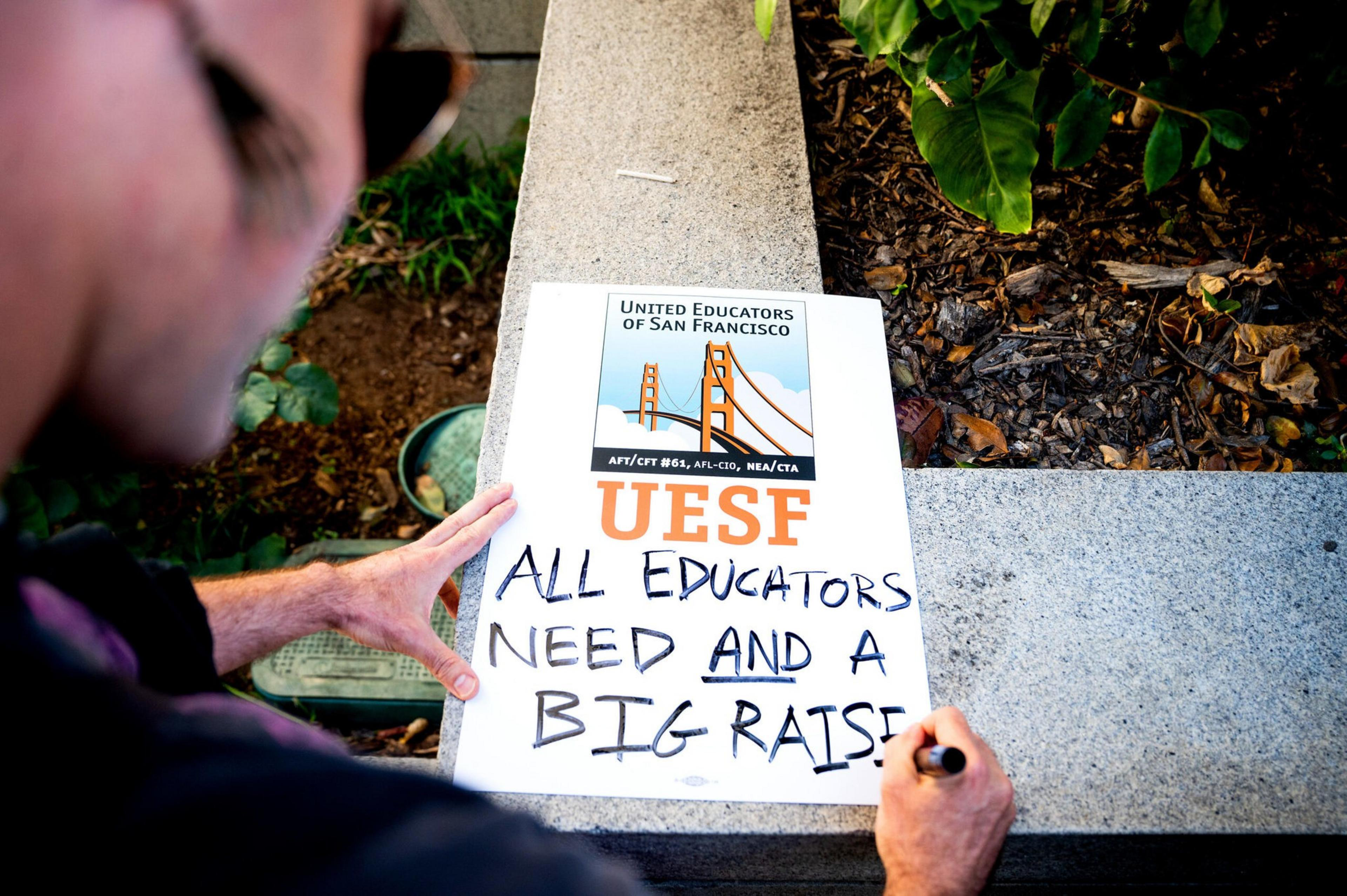 A person writes on a sign reading "UESF ALL EDUCATORS NEED AND A BIG RAISE!" with a United Educators of San Francisco logo above.