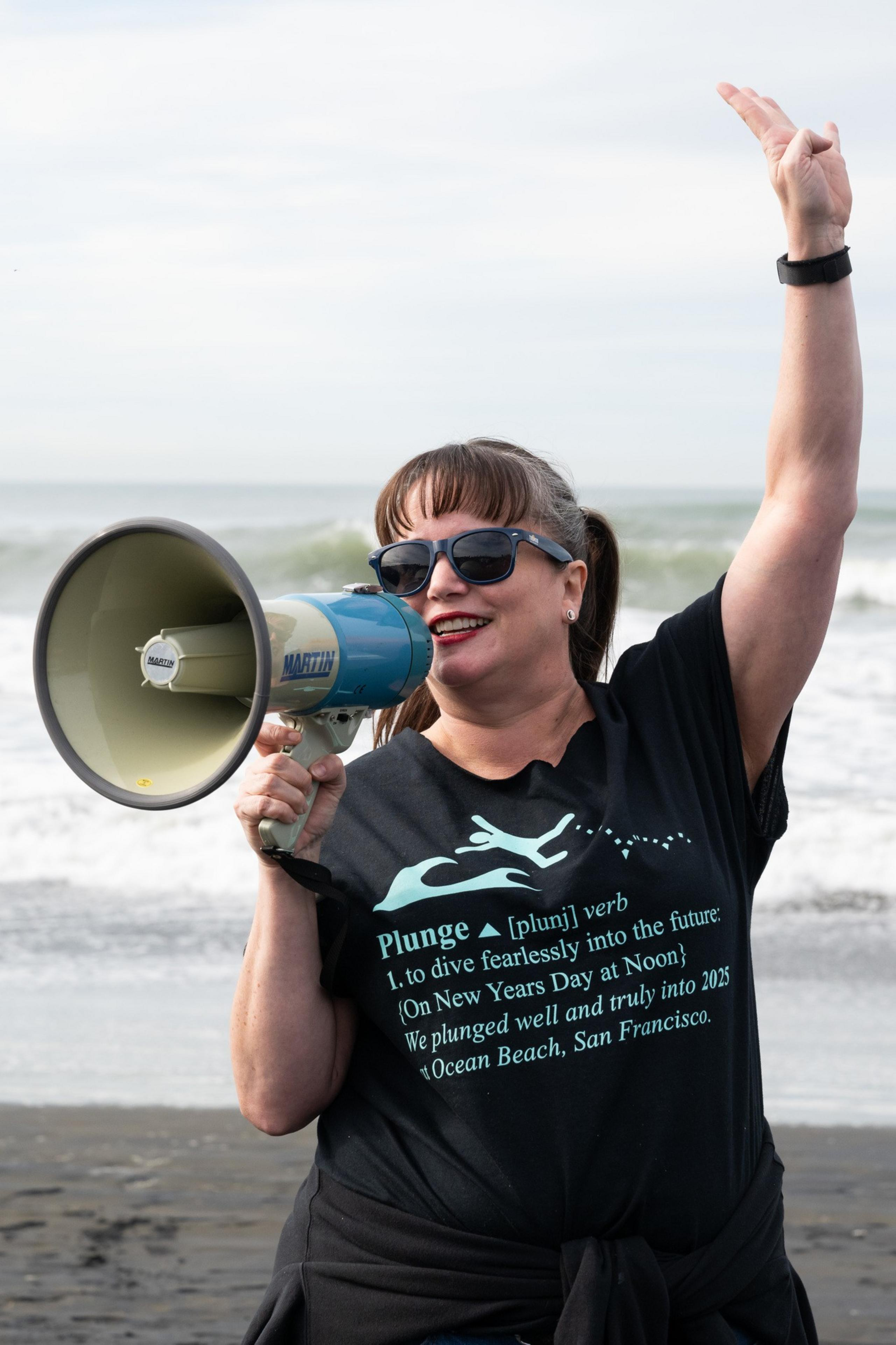 A woman in sunglasses and a black shirt holds a megaphone and raises one arm. The shirt reads &quot;Plunge: verb 1. to dive fearlessly&quot; with ocean waves behind her.