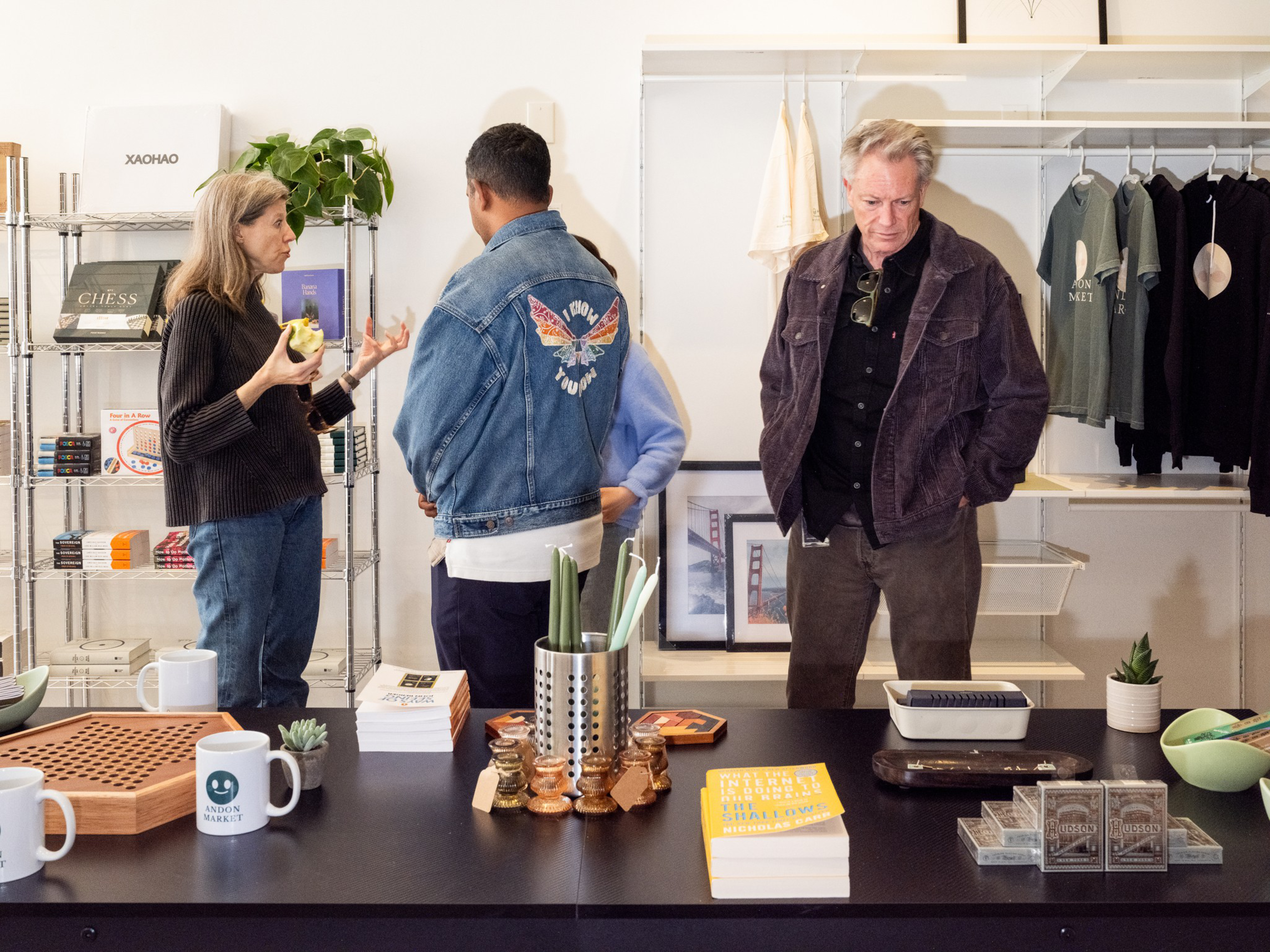 Three people stand inside a store; a woman talks holding an apple, a man in a denim jacket faces away, and an older man browses a counter with books and mugs.