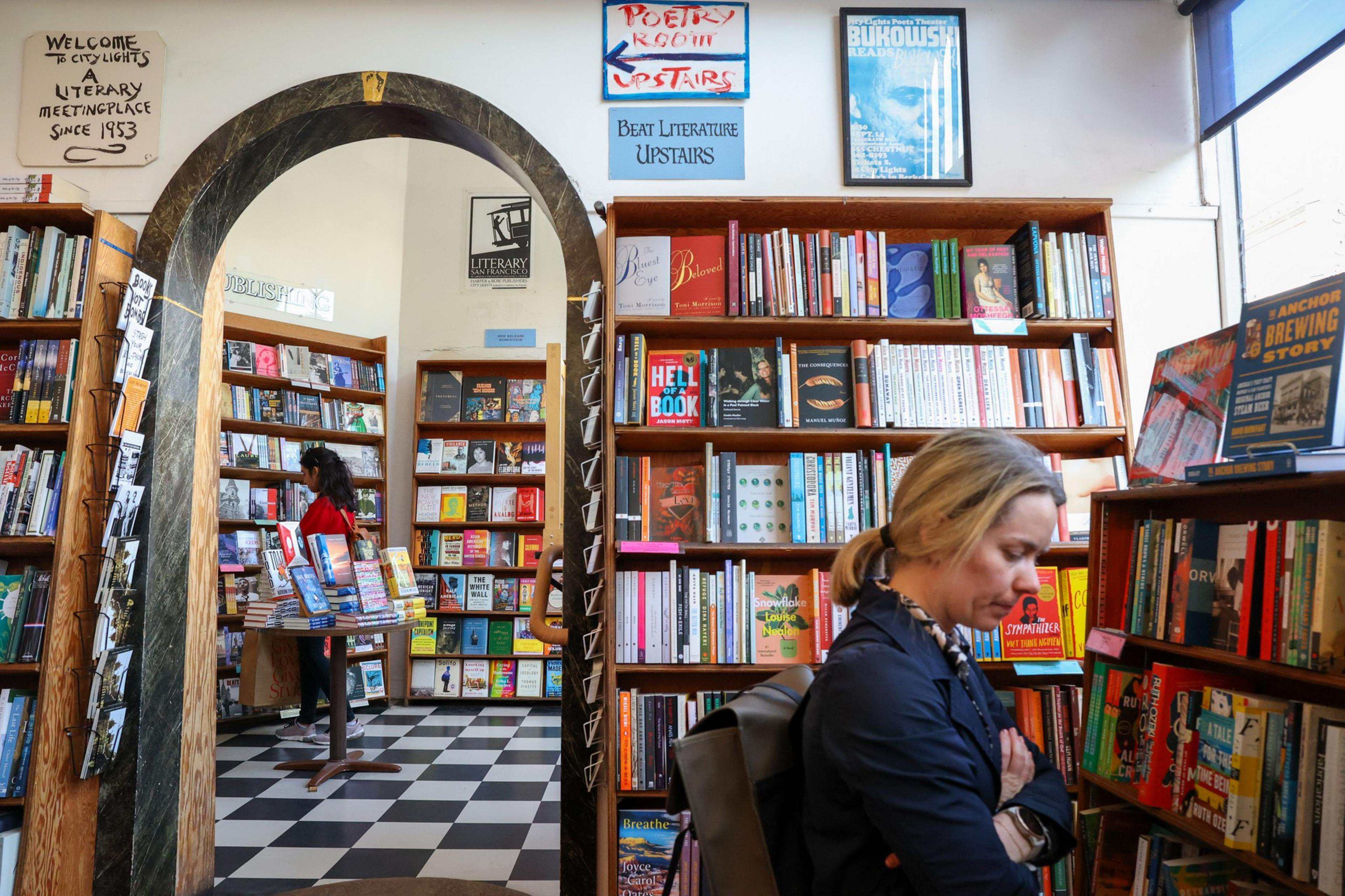 A bookstore features wooden shelves packed with books, a black-and-white tiled floor, and a woman browsing near large windows letting in daylight.