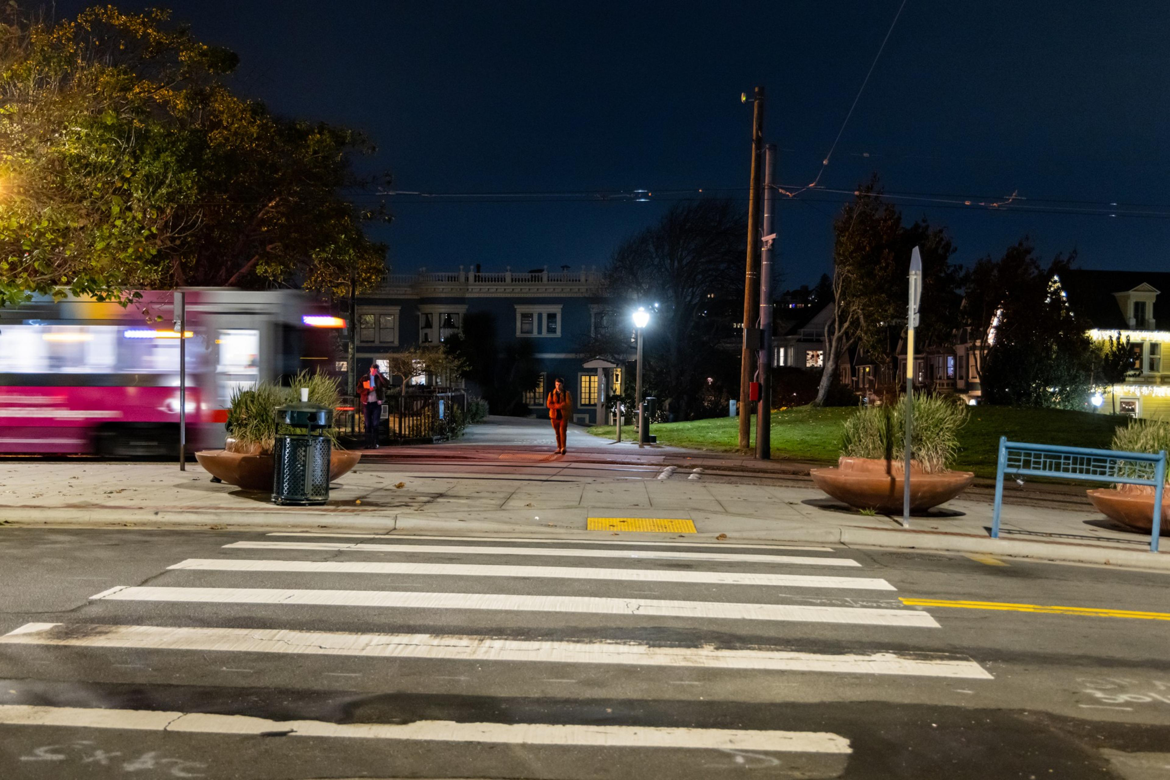A blurry pink and white tram passes on a street at night. A person walks on the sidewalk under streetlights, with trees and houses in the background.
