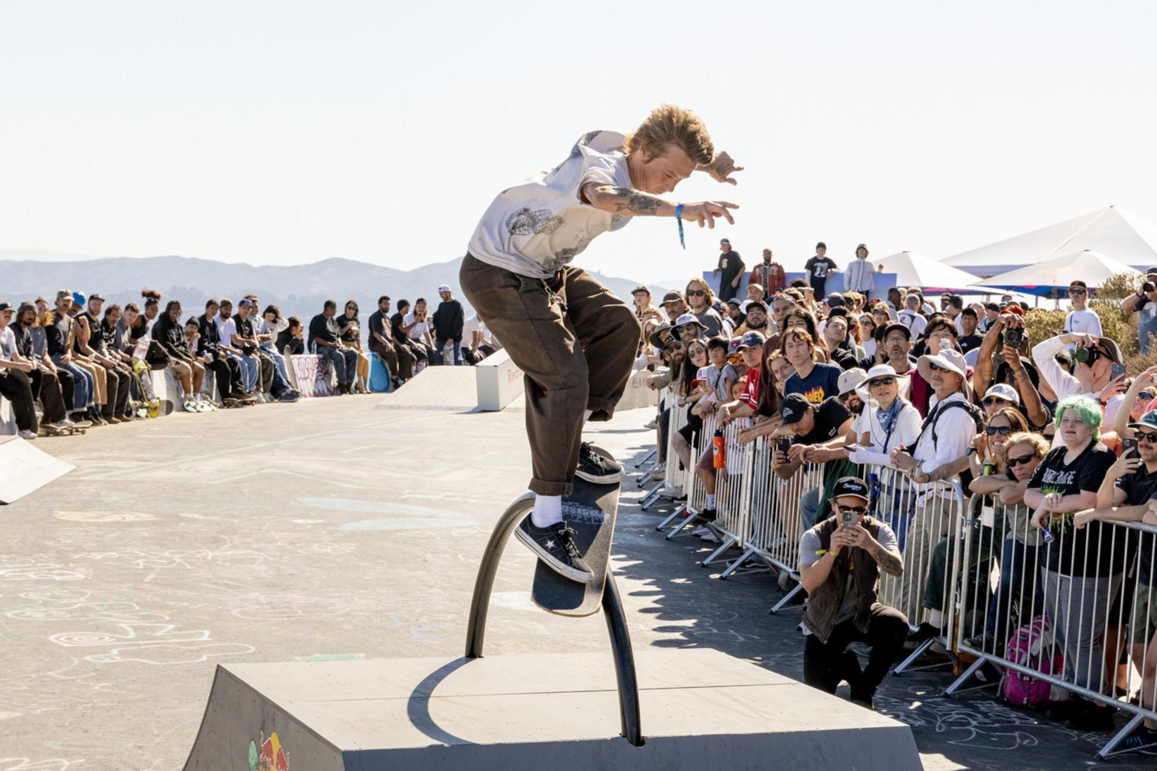 A skateboarder performs a trick on a curved rail ramp, surrounded by a large crowd of spectators. The ramp displays Red Bull branding.