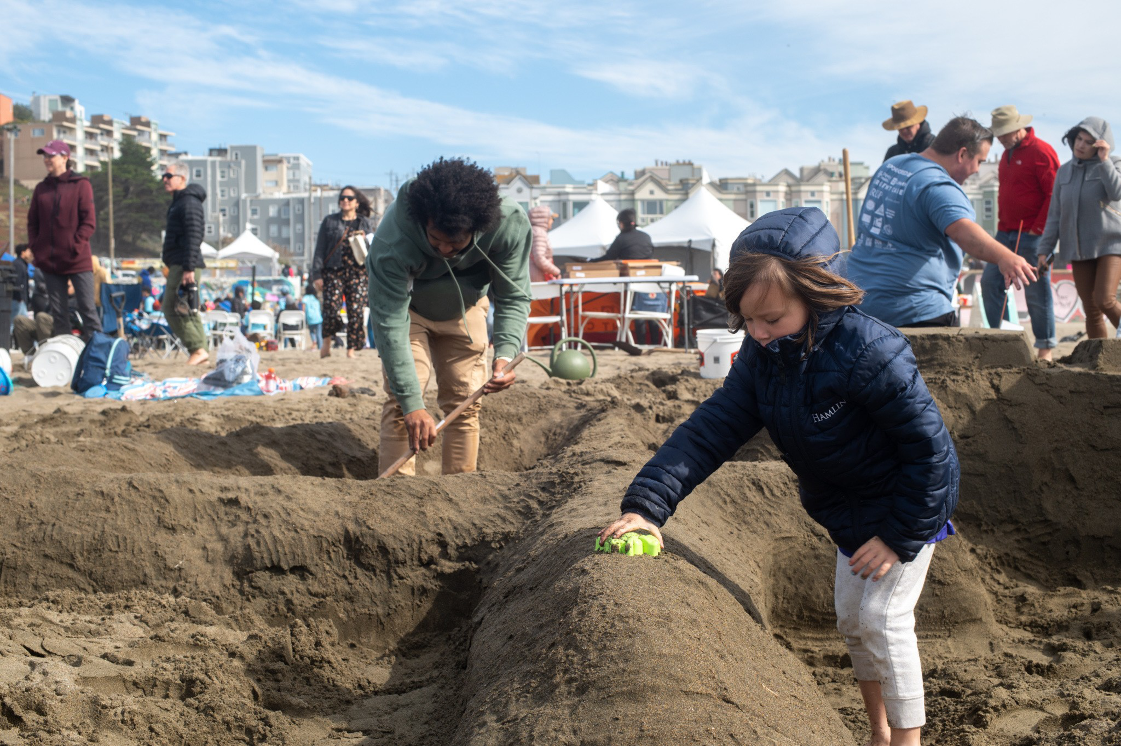 A child and an adult dig and shape large sand structures on a crowded beach with tents and buildings in the background.