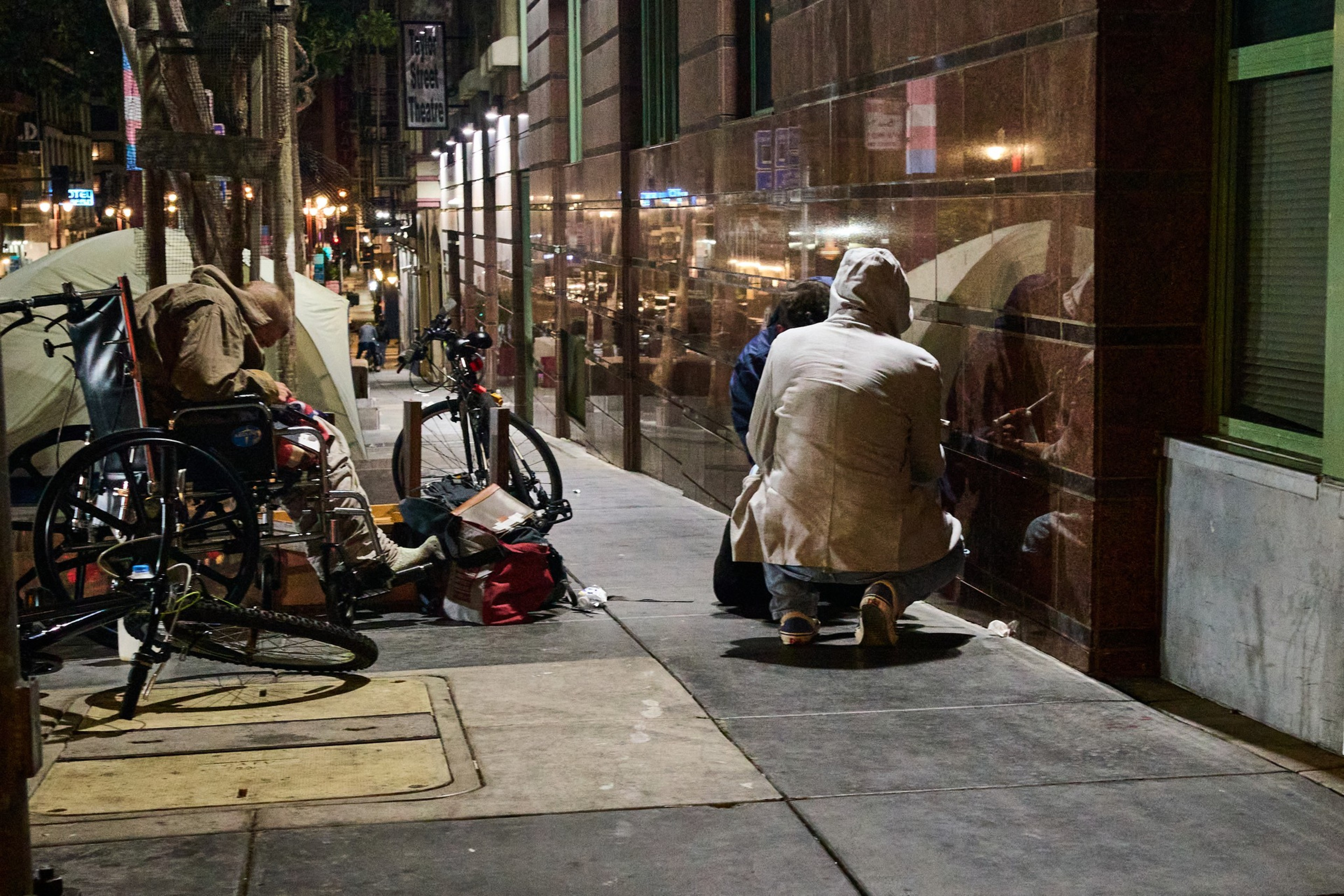 A nighttime street scene shows two people sitting on a sidewalk by a building. A wheelchair and bicycles are nearby, with tents and streetlights in the background.