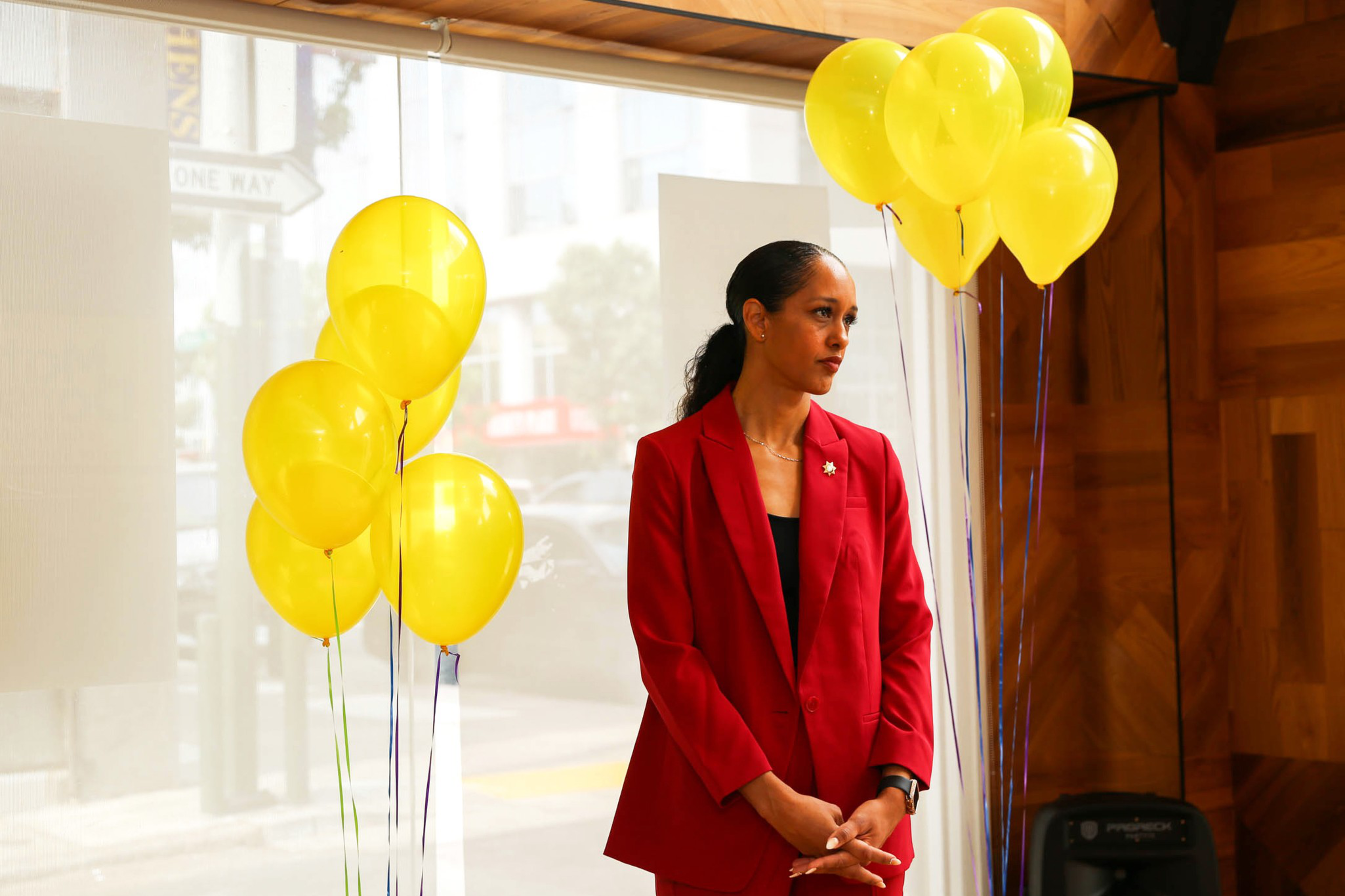 A woman in a red suit stands between two bunches of yellow balloons in a room with wooden walls and sunlight coming through large windows.