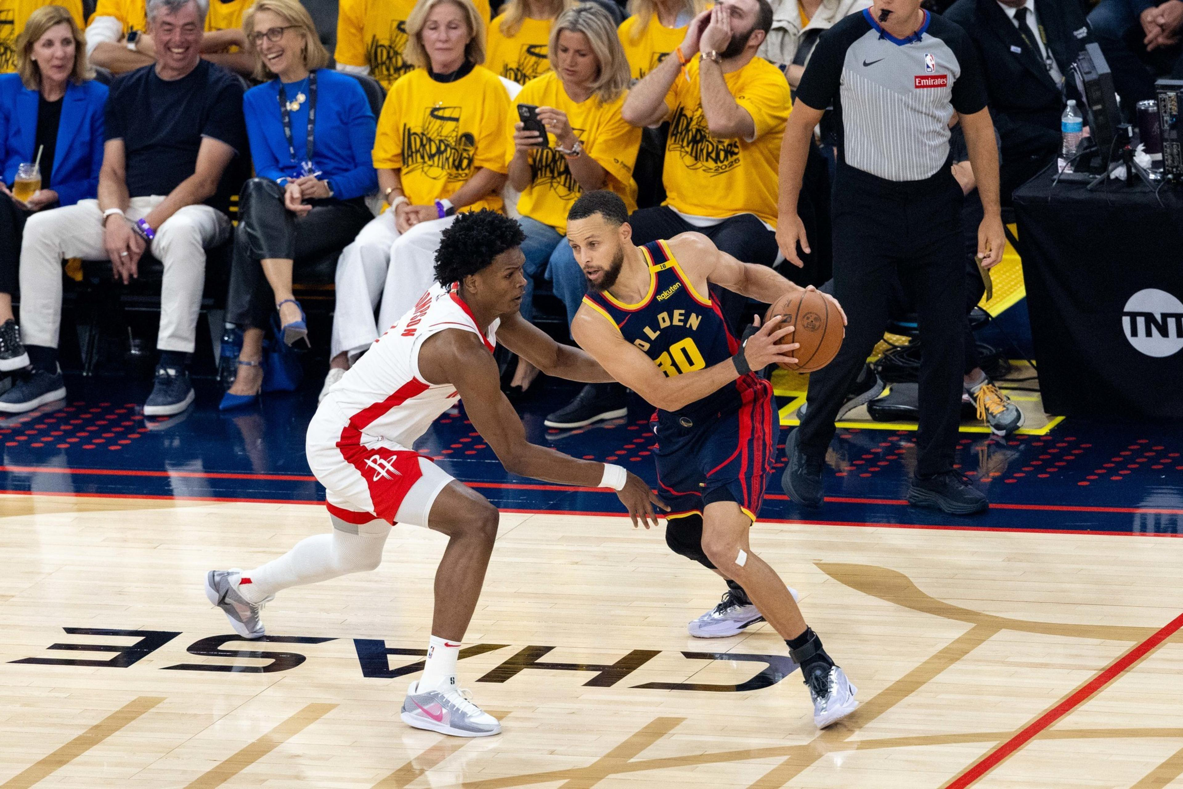 Two basketball players are on the court, one dribbling the ball, while the crowd watches intently. A referee stands nearby, and fans wear team shirts.