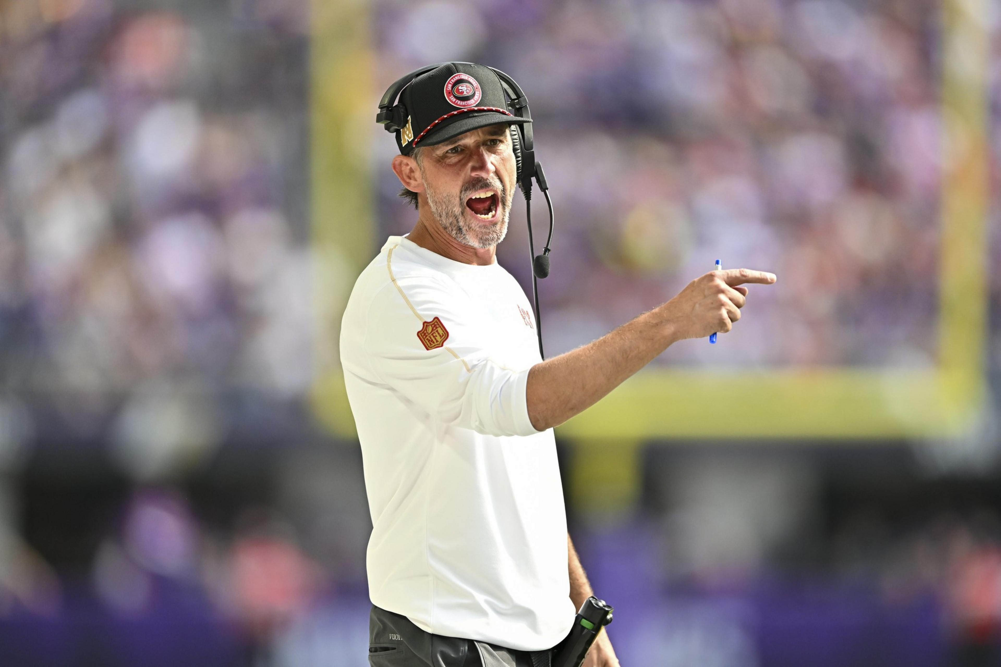 A man wearing a headset and cap is passionately gesturing and pointing with a pen. He is dressed in a white shirt, possibly during a sports event.