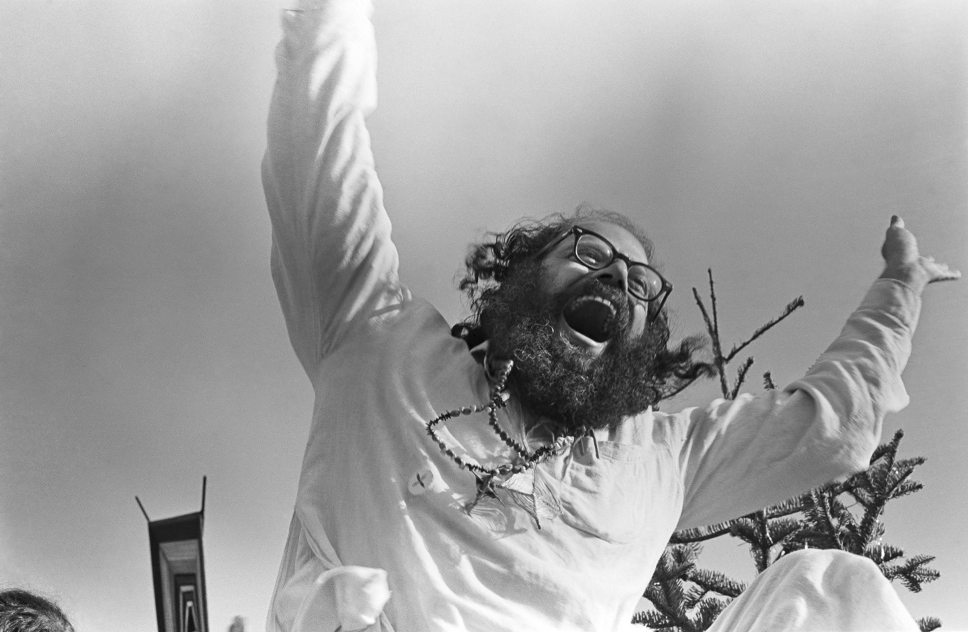 A bearded man with glasses joyfully throws his arms up against a clear sky. He's wearing a light shirt and a beaded necklace, and appears exuberant and lively.