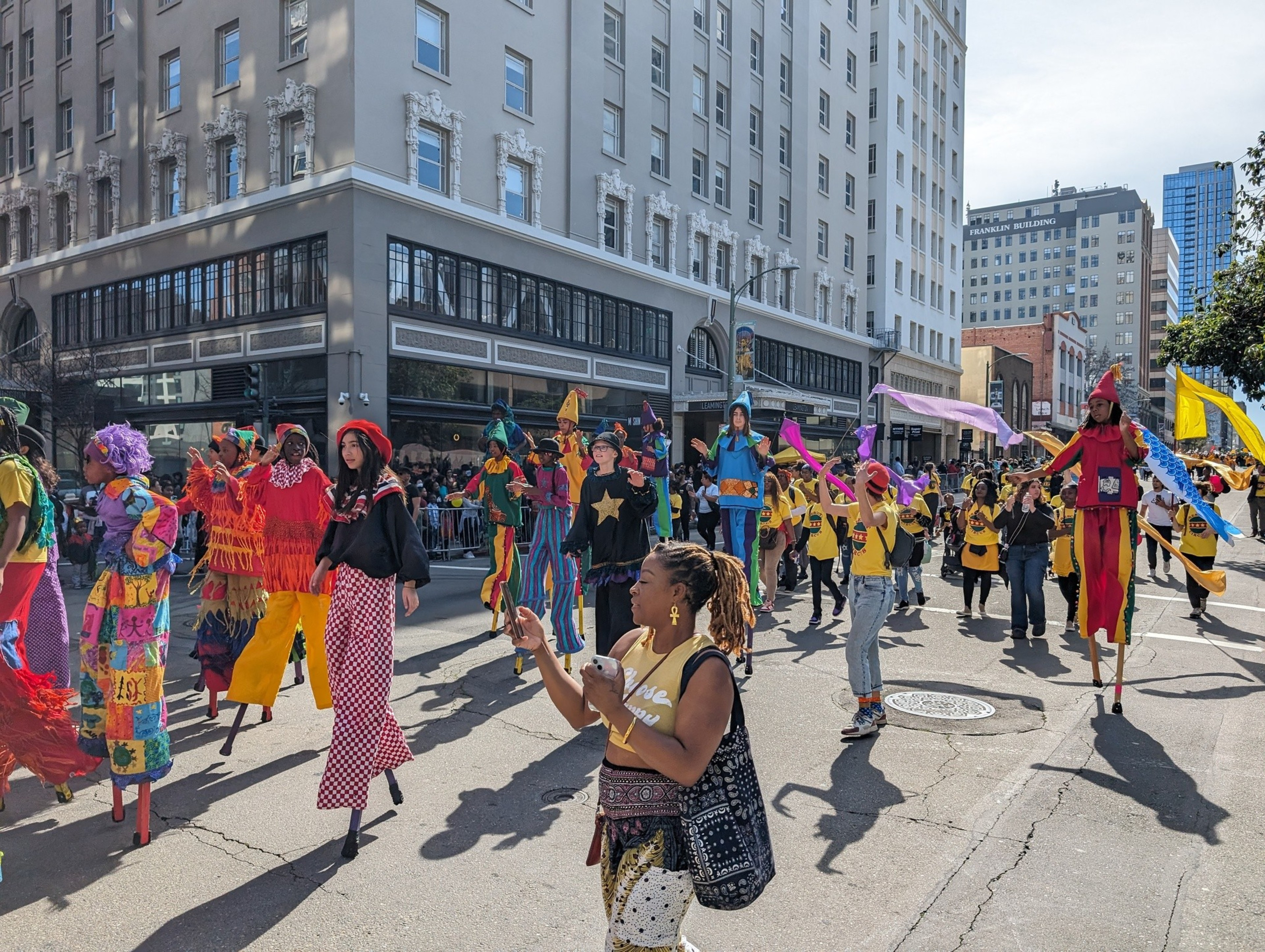 Several young people gracefully walk on stilts along a parade route lined with cheering visitors.