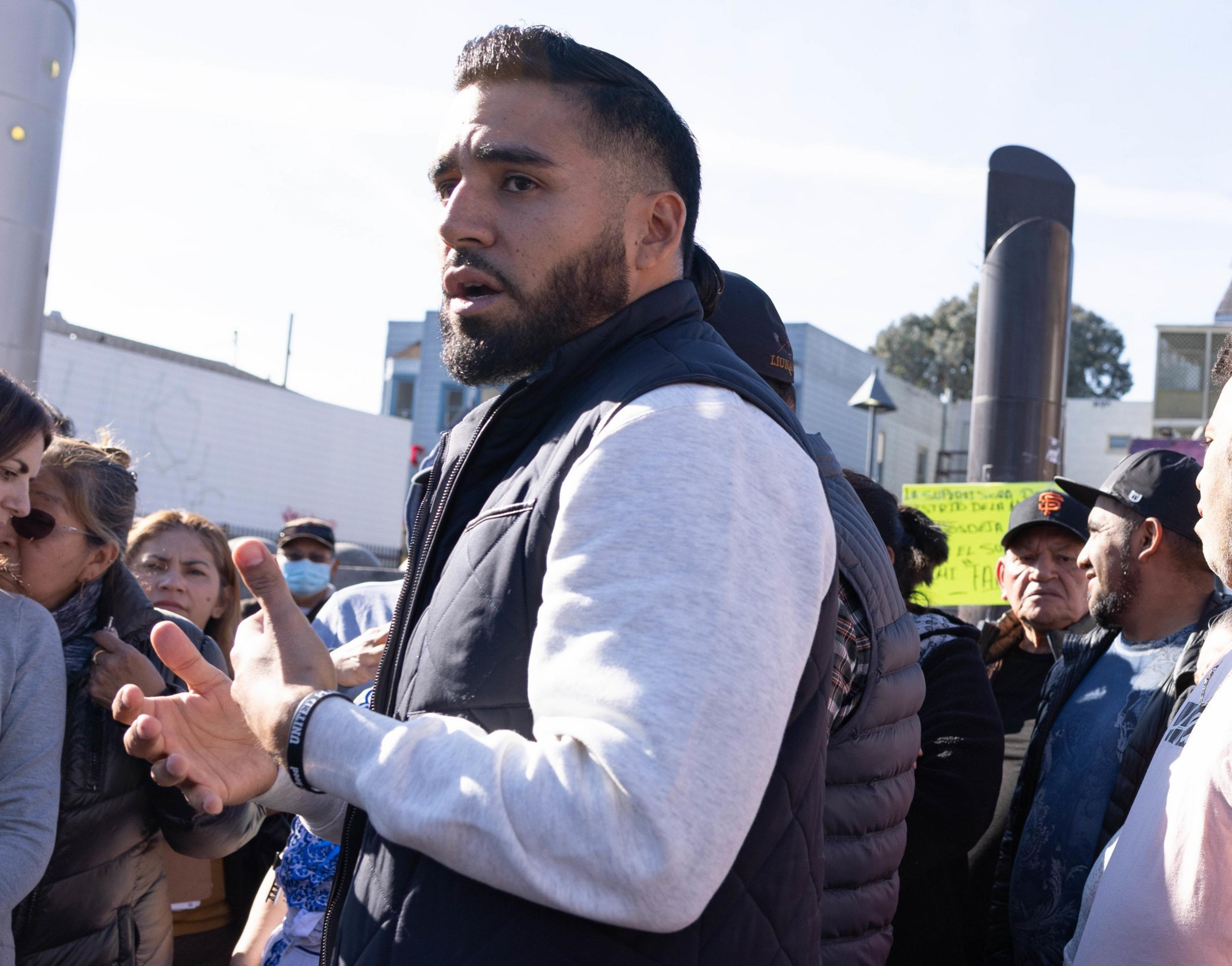 A man gestures mid-speech in a gathered crowd, looking concerned or expressive.