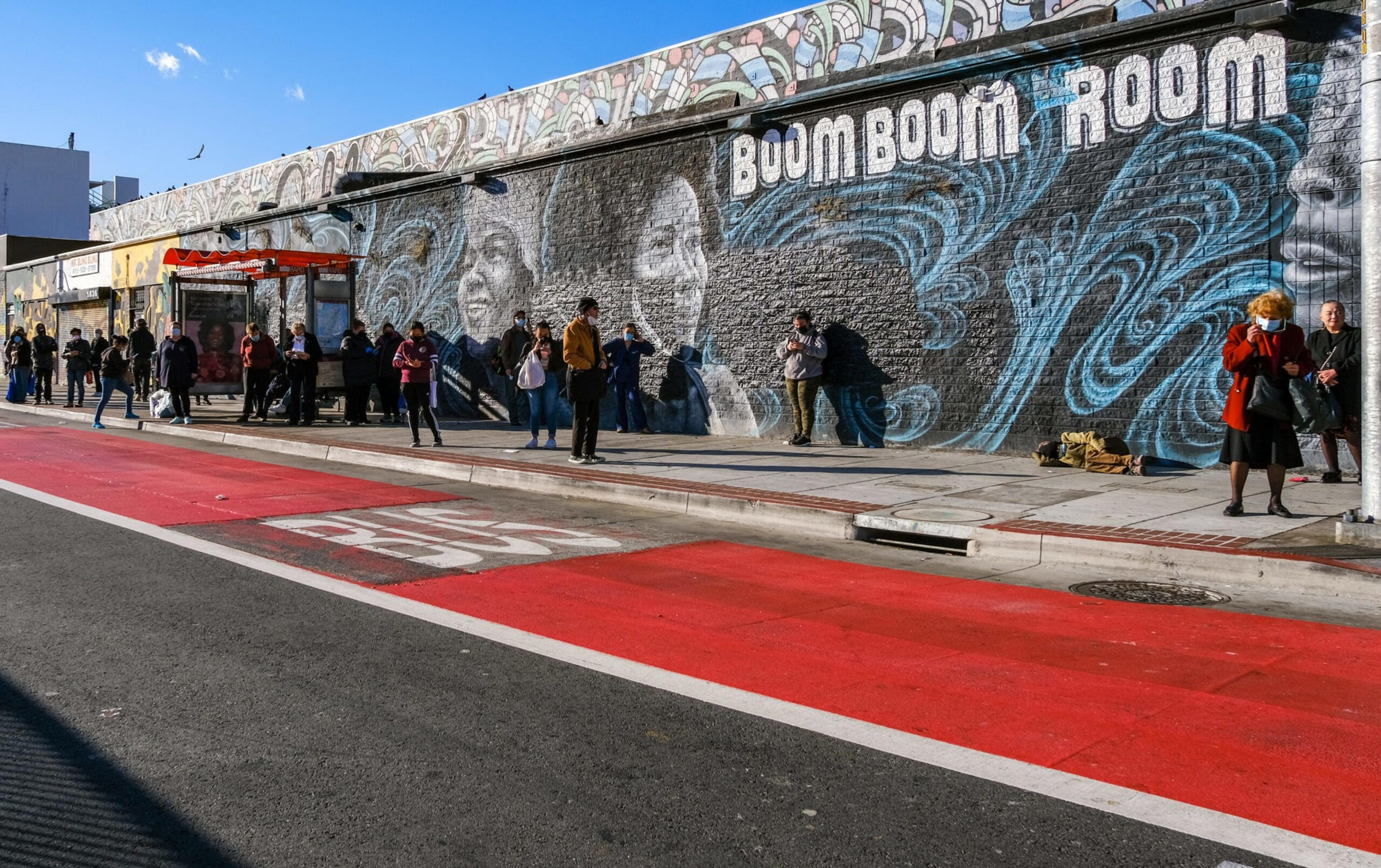 A group of people wait at a bus stop beside a colorful mural with three smiling faces and the words "Boom Boom Room."