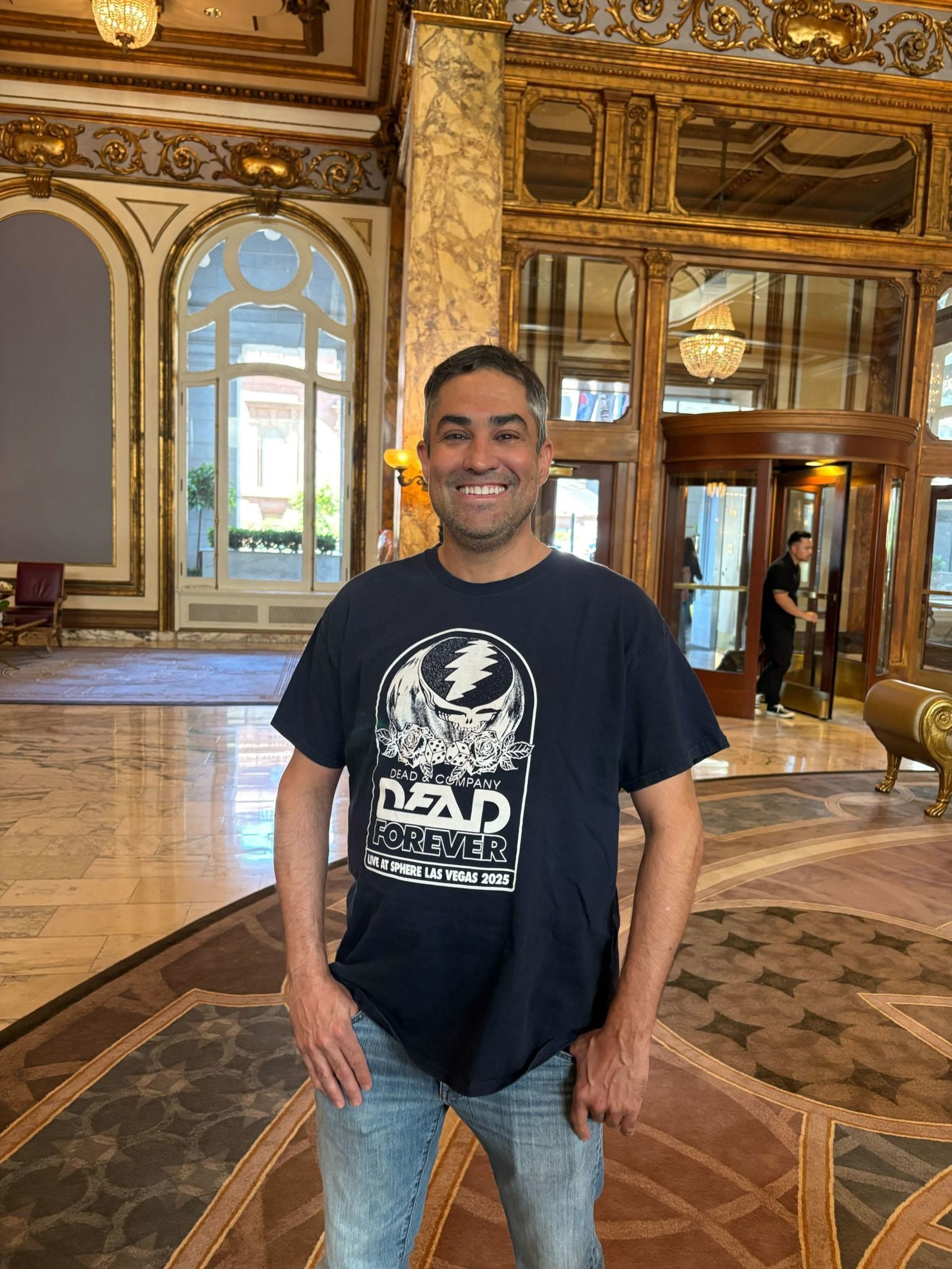 A man smiles while standing in a grand, ornate room with marble floors, gold-trimmed mirrors, and a revolving door in the background.