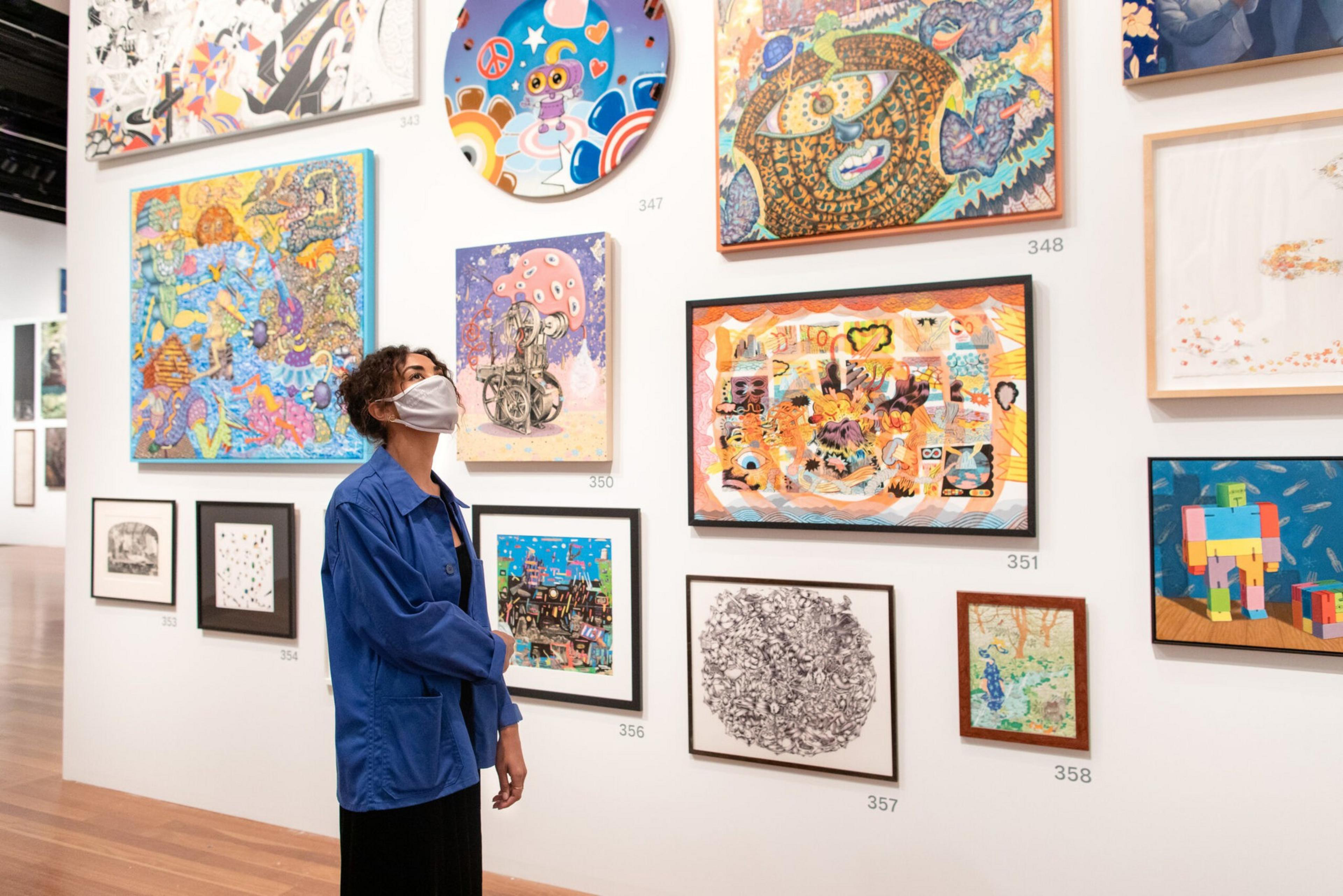 A person wearing a mask and blue jacket observes a wall filled with colorful, abstract, and detailed artworks in a gallery.