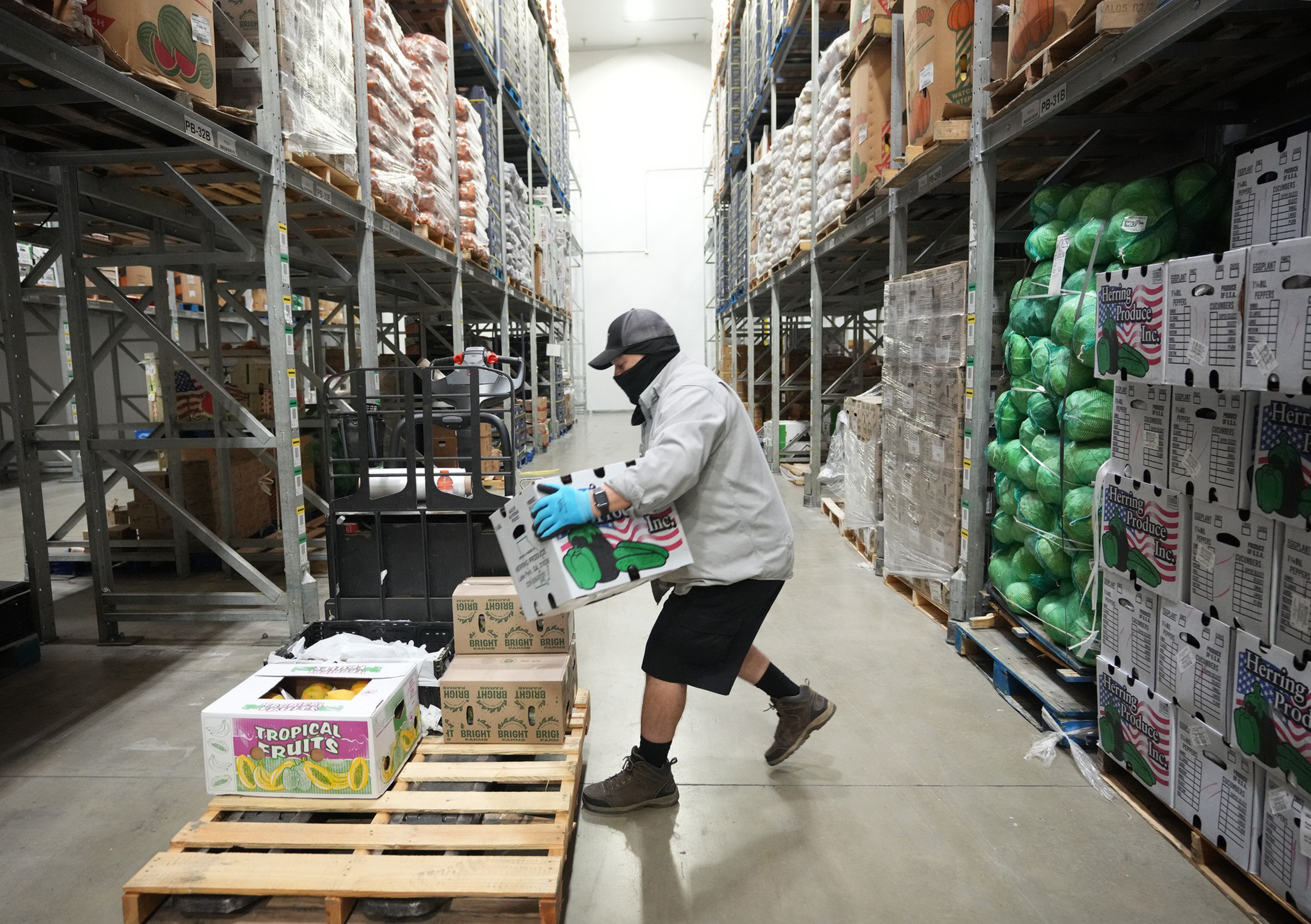 A person wearing a cap, mask, gloves, and shorts lifts a box in a warehouse aisle stacked with boxes and produce on metal shelves.