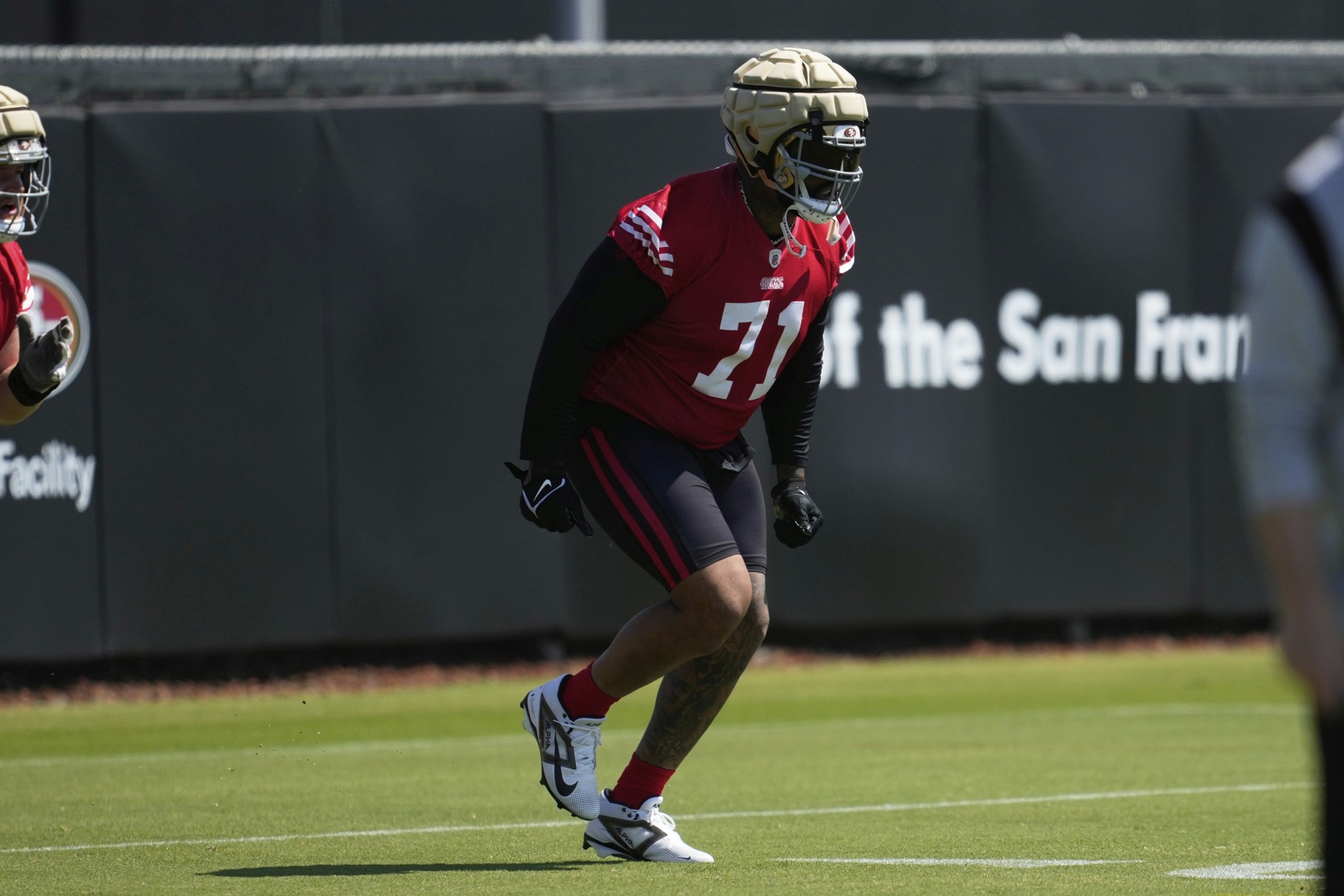 A football player wearing a red jersey with the number 71, a padded helmet, and black leggings, is running on a grassy field during practice.