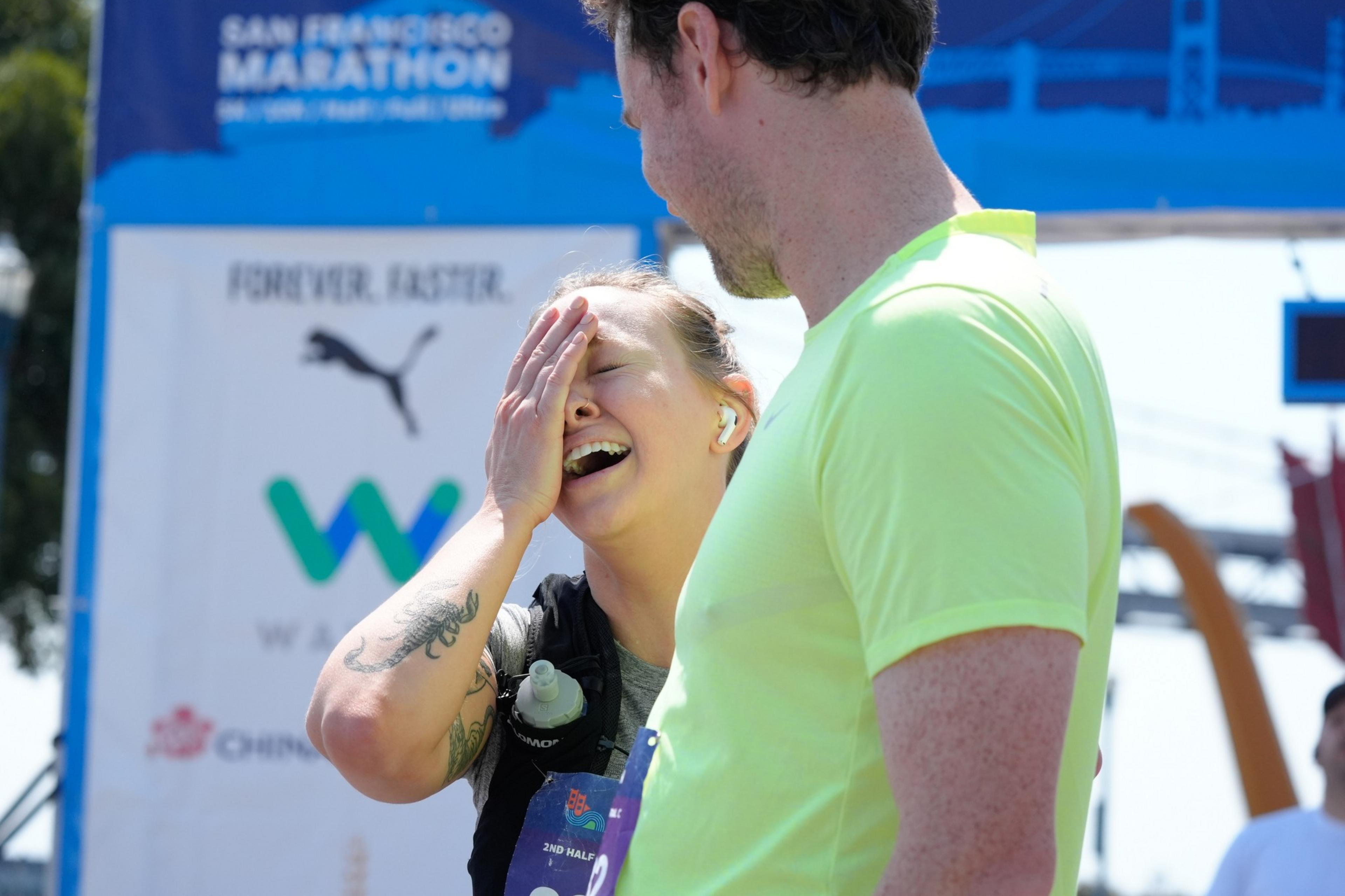 A woman laughs while covering her face, standing beside a man in a neon yellow shirt at the San Francisco Marathon finish line, with a blue marathon banner in the background.