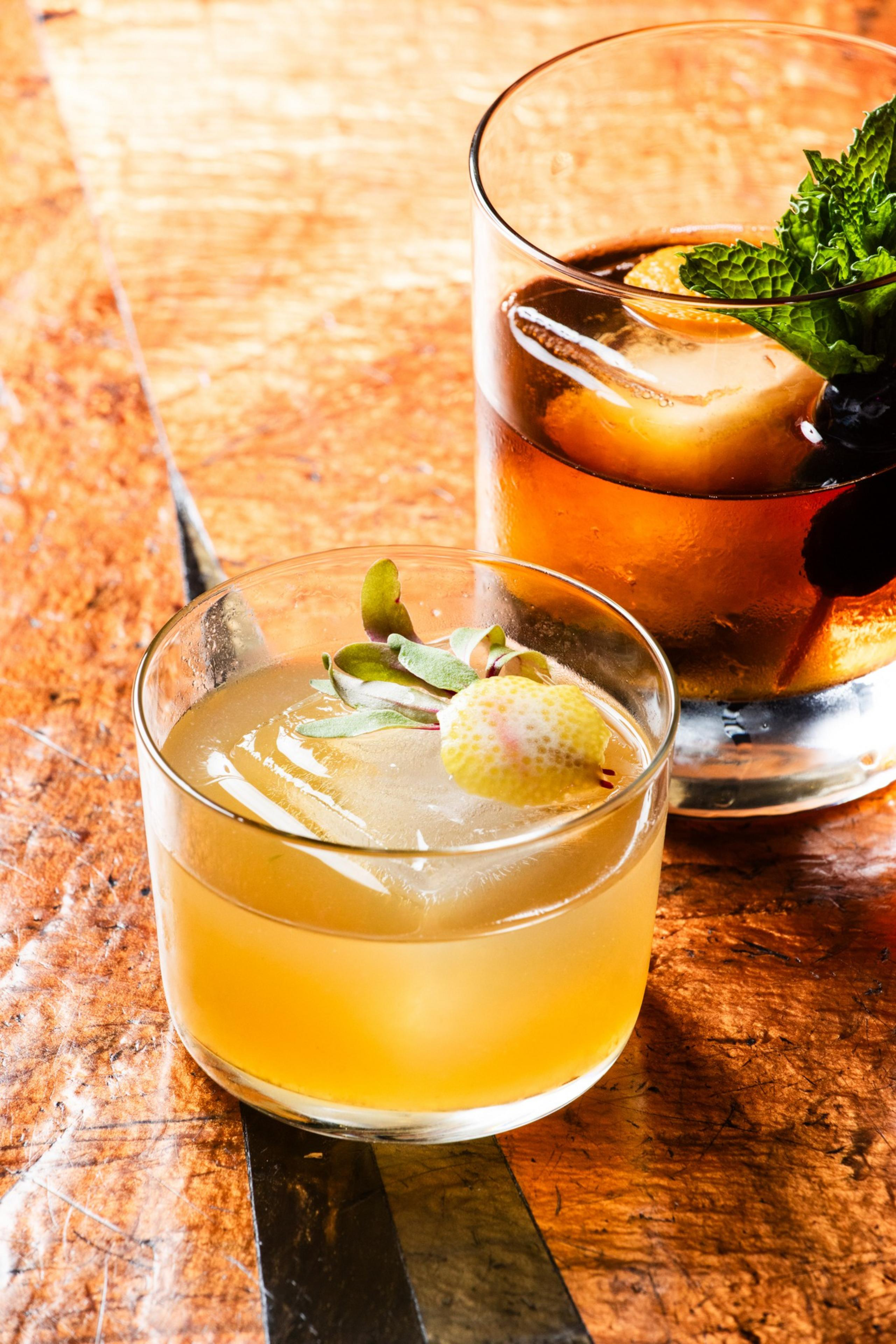 Two cocktails sit on a wooden table: one light-colored with a lemon and herb garnish, and another darker drink with ice, mint, and a garnish.