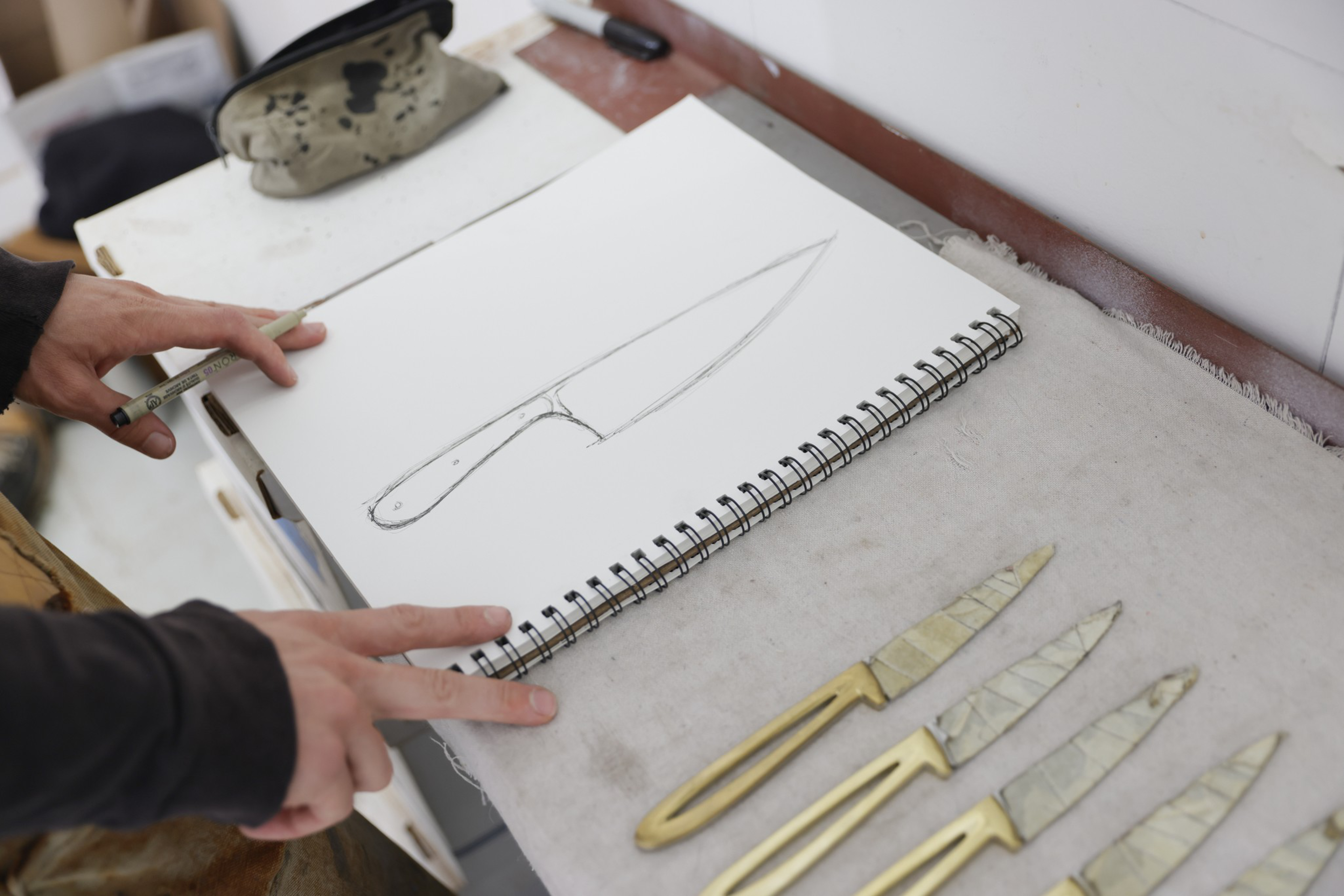 A hand is sketching a large knife on a spiral-bound sketchpad, with four brass-handled knives arranged on the table beside it.
